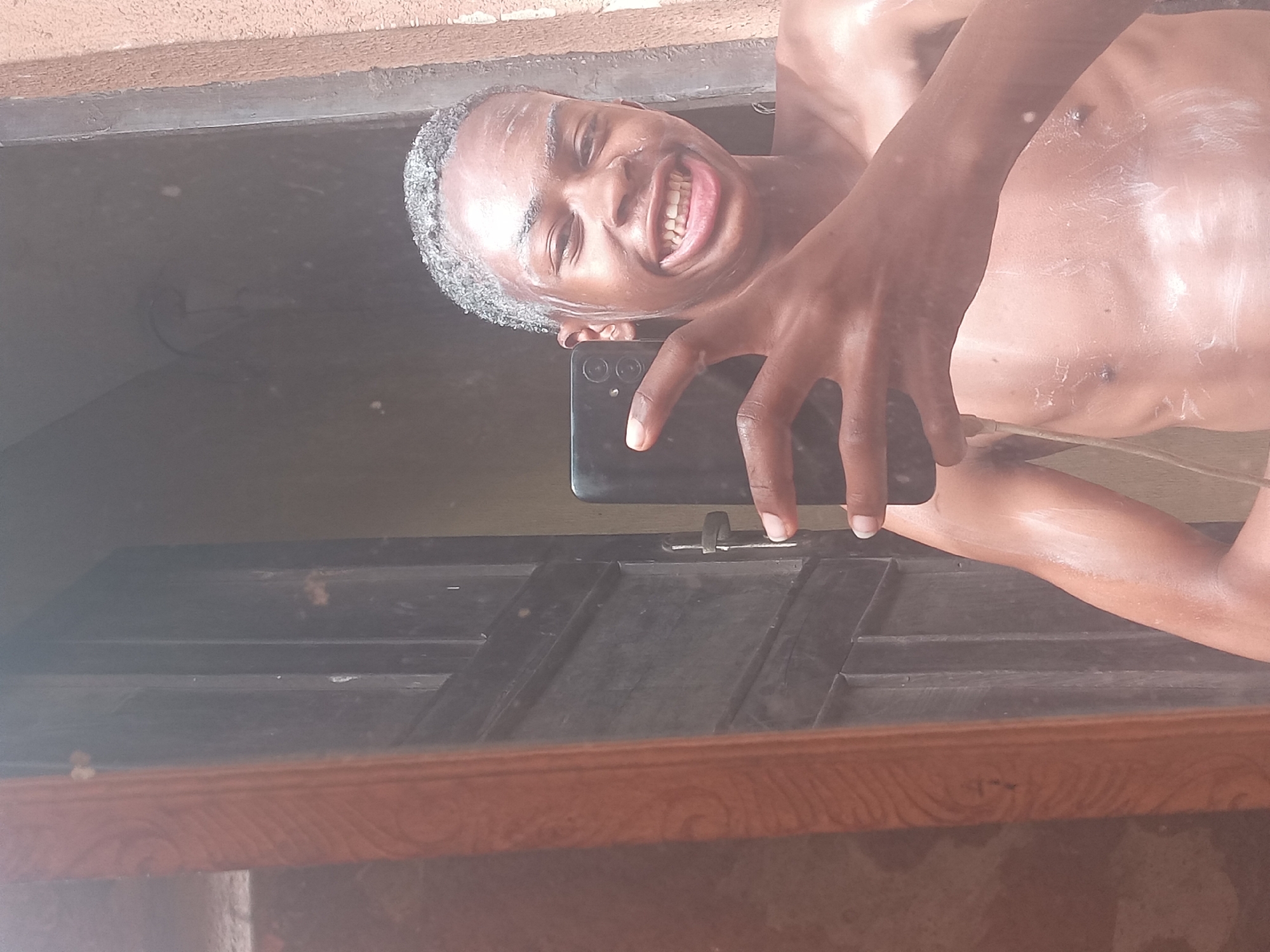 A person with soap on their body and hair is smiling while holding a black phone in front of them, likely taking a selfie in a mirror. The reflection shows a dark-stained wooden surface with a light brown patterned edge. The location is Onitsha, Nigeria.