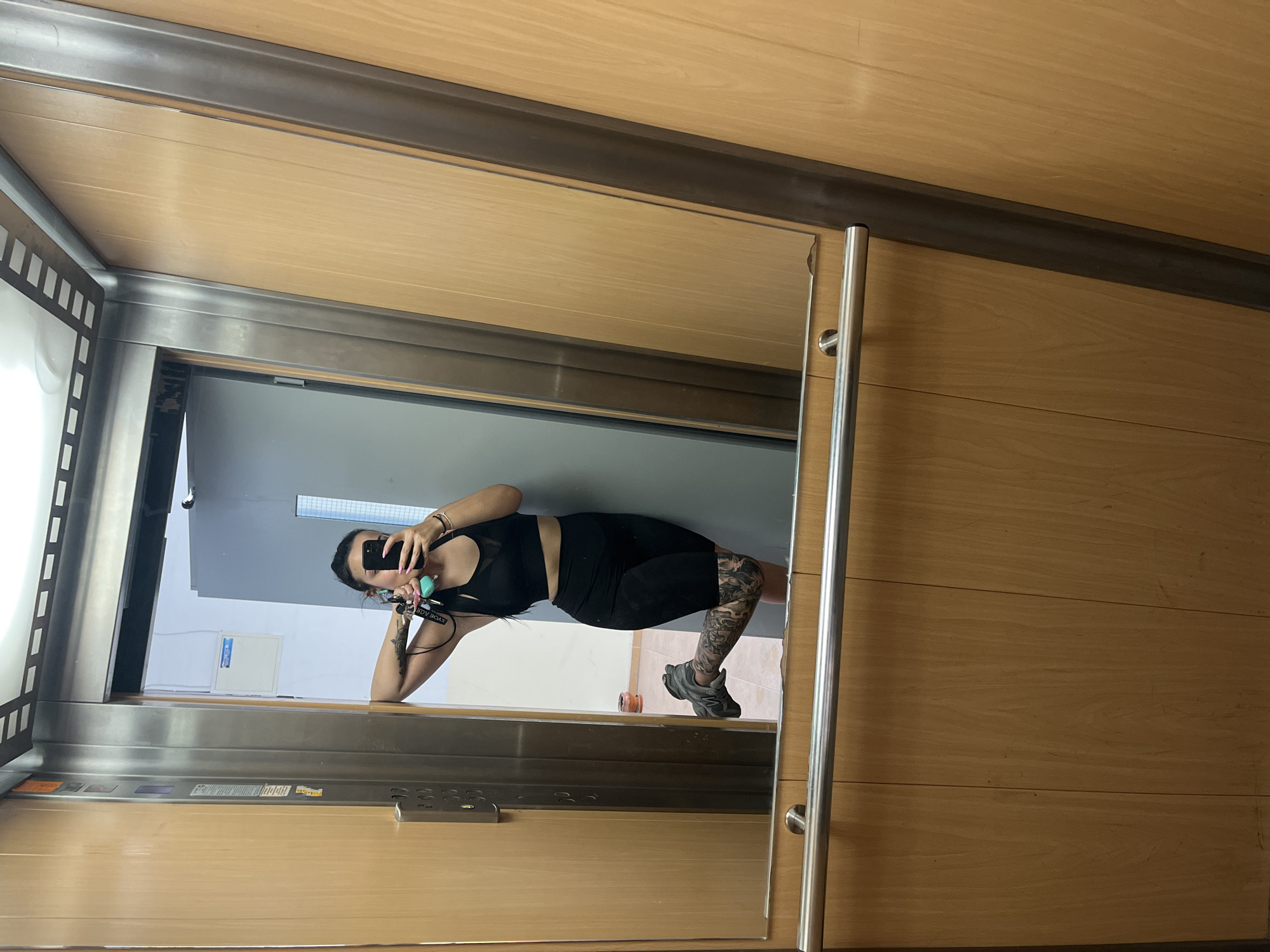 A person is captured in a mirror selfie inside an elevator, appearing to be in Veliko Tarnovo, Bulgaria. The image is taken from a slightly low angle, showing the person holding a smartphone to obscure their face.
The subject is wearing a black athletic crop top and black cycling shorts or fitted shorts, paired with grey sneakers. Their left leg is extensively covered with black and grey tattoos, visible from the thigh down to the calf. A smaller tattoo is also visible on their inner left arm/wrist. They wear a wristwatch or bracelet on their right wrist and rings on several fingers. A keychain with multiple keys and a light blue/green accessory is held in their left hand, with the words "ROYAL ROCKS" partially visible on it.
The setting is an elevator interior featuring light wooden paneling on the walls and ceiling. A large mirror covers one wall, reflecting the person and the interior of the elevator. The mirror is framed by metal accents, and a metal handrail is present on the right side of the mirror. Below the mirror, a control panel with silver buttons and an unidentifiable digital display or small screen is integrated into the metal and wood structure.
Through the reflection in the mirror, elements outside the elevator are visible, including a plain white wall, a light grey door frame, and a white rectangular box with blue labels (possibly an electrical panel) mounted on the wall. The overall lighting suggests daytime. The activity is clearly a mirror selfie, with no specific emotional expression discernible due to the phone covering the subject's face.