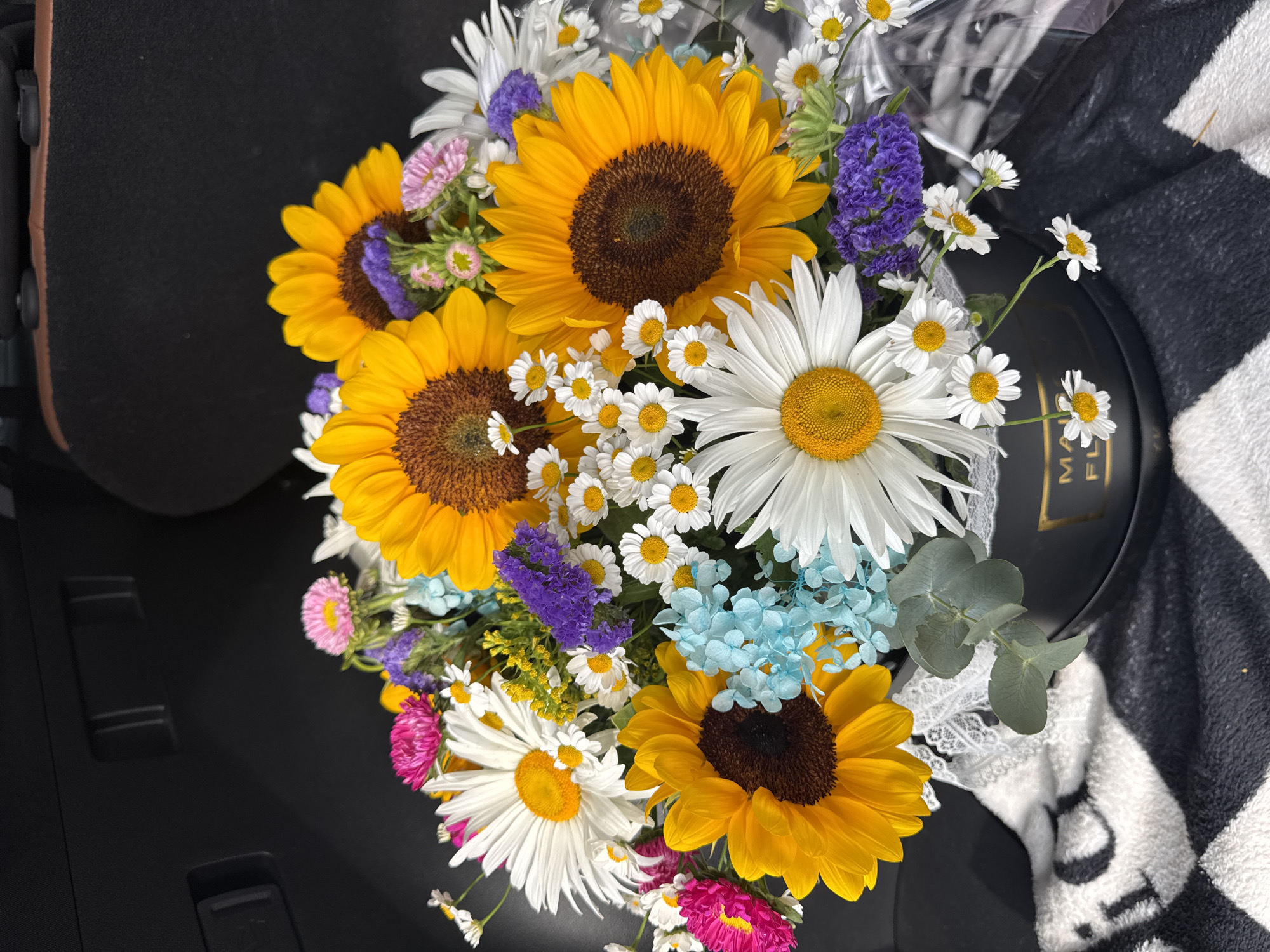 A close-up, overhead shot captures a vibrant bouquet of sunflowers, daisies, pink asters, blue hydrangeas, and purple statice flowers arranged in a black circular gift box. The box, adorned with a gold emblem that reads "MAISON FILS," sits on a black and white patterned blanket. The background consists of the dark, textured interior of a vehicle, possibly a car trunk, with visible dark gray and brown upholstery. The lighting suggests an outdoor setting during the daytime, casting natural light on the colorful floral arrangement. The image is sharp and well-lit, highlighting the intricate details of each flower and the textures of the surrounding objects. The overall scene evokes a sense of freshness and celebration. There are no people visible in the image. The location is not specified as Almaty, Kazakhstan.