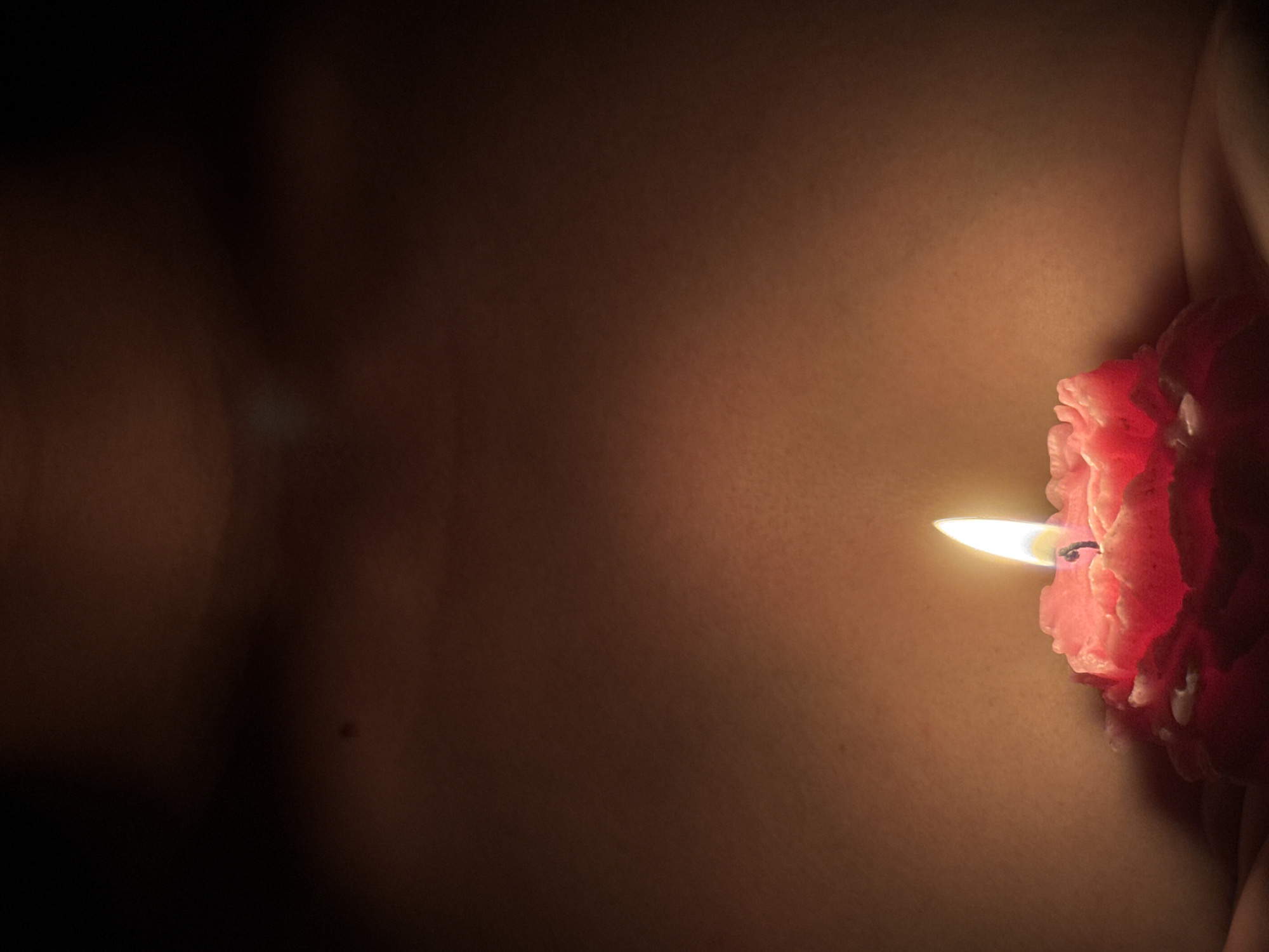 A lit pink candle with a flame sits in the center of the frame, casting a warm glow on the surrounding skin. The candle is shaped like a flower and appears to be held in hands. The background is a dark, softly lit expanse of skin, possibly a torso, with subtle shadows and highlights. A small, dark mole is visible on the upper left portion of the skin. The overall impression is intimate and moody. The location in Chaikovsky, Russia, cannot be determined from the image.