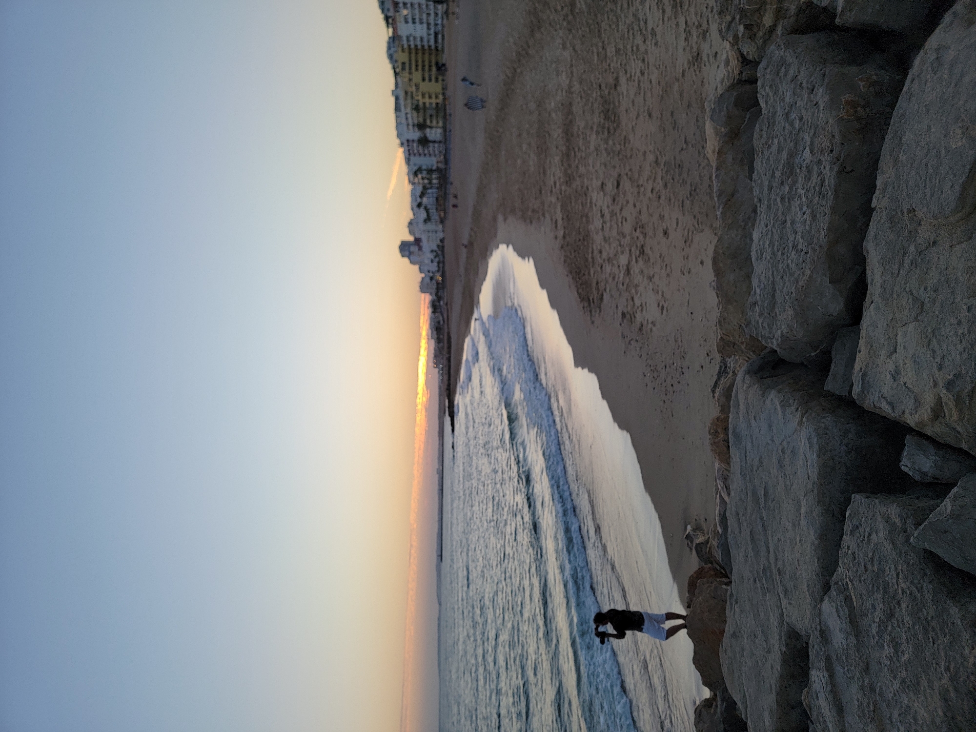 A vertical, eye-level, outdoor, medium shot captures a tranquil beach scene in Quarteira, Portugal, at dusk. The sky is a gradient of soft blues and oranges, transitioning to a pale yellow near the horizon where the sun has just set. The ocean, to the left, is a deep blue with gentle waves lapping at the shore. A lone figure, silhouetted against the water, stands on a rocky outcrop, holding a camera to their eye, seemingly taking a photograph. They are wearing a dark shirt and white shorts. The sandy beach curves to the right, with a few distant figures walking along it. Along the coastline, a line of white and yellow buildings with multiple stories forms the backdrop of the town, interspersed with palm trees. In the foreground, large, rough grey rocks are prominently featured, adding texture and a sense of scale to the scene. The overall atmosphere is peaceful and serene.
