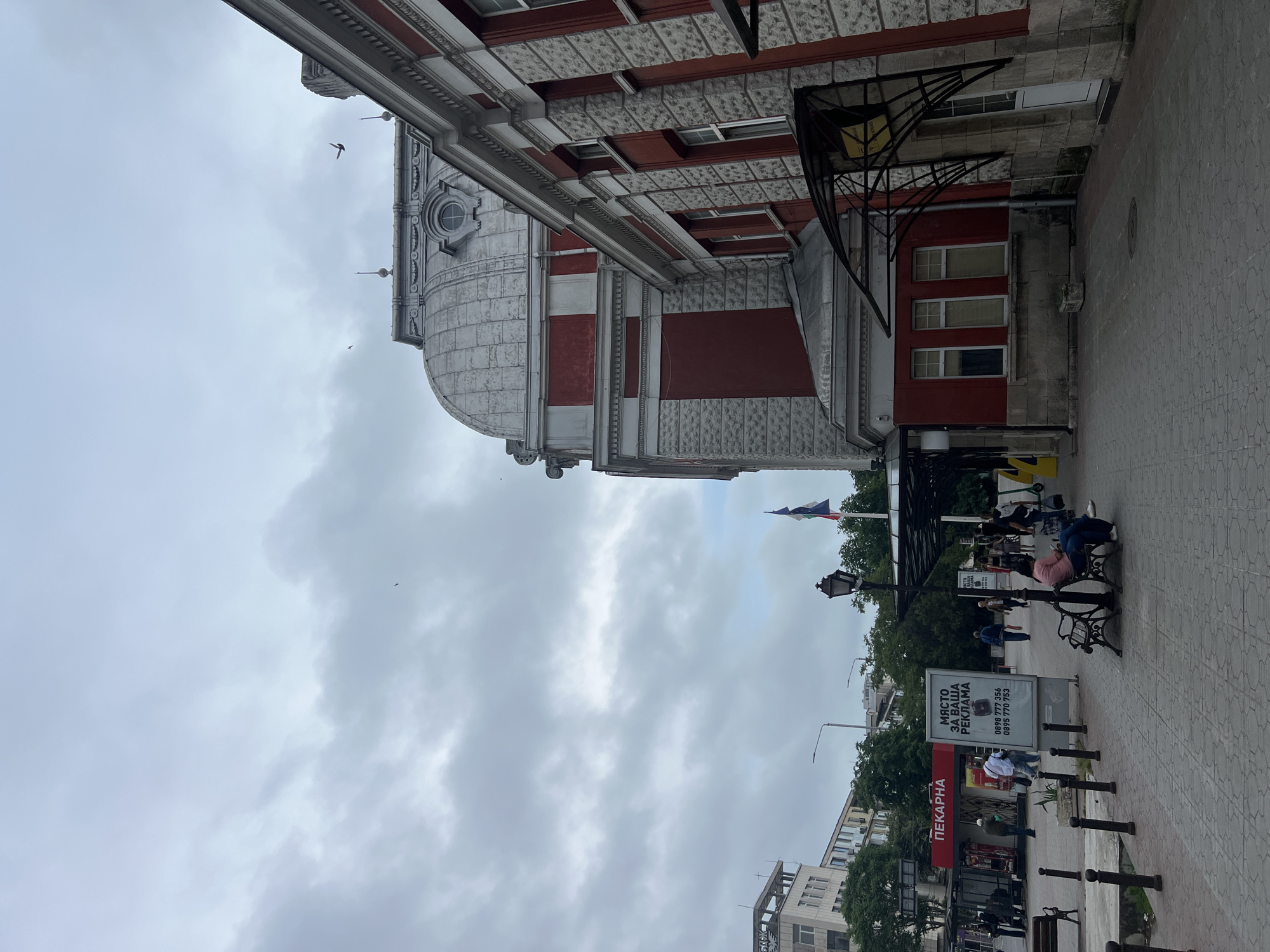 The image captures an overcast daytime street scene in Varna, Bulgaria, featuring a mix of historical and urban elements.
On the left, a grand, multi-story building dominates the frame with its distinctive architecture. It showcases a large, grey domed section at its top, adorned with a circular window. The facade is detailed with alternating bands of reddish-brown and textured grey stone, complemented by intricate white cornices, window frames, and decorative elements.
The right side of the street features a pedestrian area with dark, patterned paving. Black, ornate metal benches and lampposts are interspersed along the sidewalk, alongside a line of black bollards. Further down the street, glimpses of more modern buildings and lush green trees are visible. A red shop sign, prominently displayed, reads "ПЕКАРНА" (Bakery). An A-frame advertising board on the sidewalk presents the text "МЯСТО ЗА ВАША РЕКЛАМА" (Place for Your Advertisement), followed by two phone numbers: "0898 777 356" and "0895 770 755".
Several people are seen engaging in casual activity on the street; some are seated on the benches, while others walk along the sidewalk. The sky is largely overcast with soft grey clouds, suggesting a cloudy day without direct sunlight. In the distance, a Bulgarian flag (white, green, and red stripes) flies on a pole, providing a clear geographical cue for Varna, Bulgaria. Two small birds are visible in flight against the sky, adding a dynamic touch to the otherwise calm urban landscape.