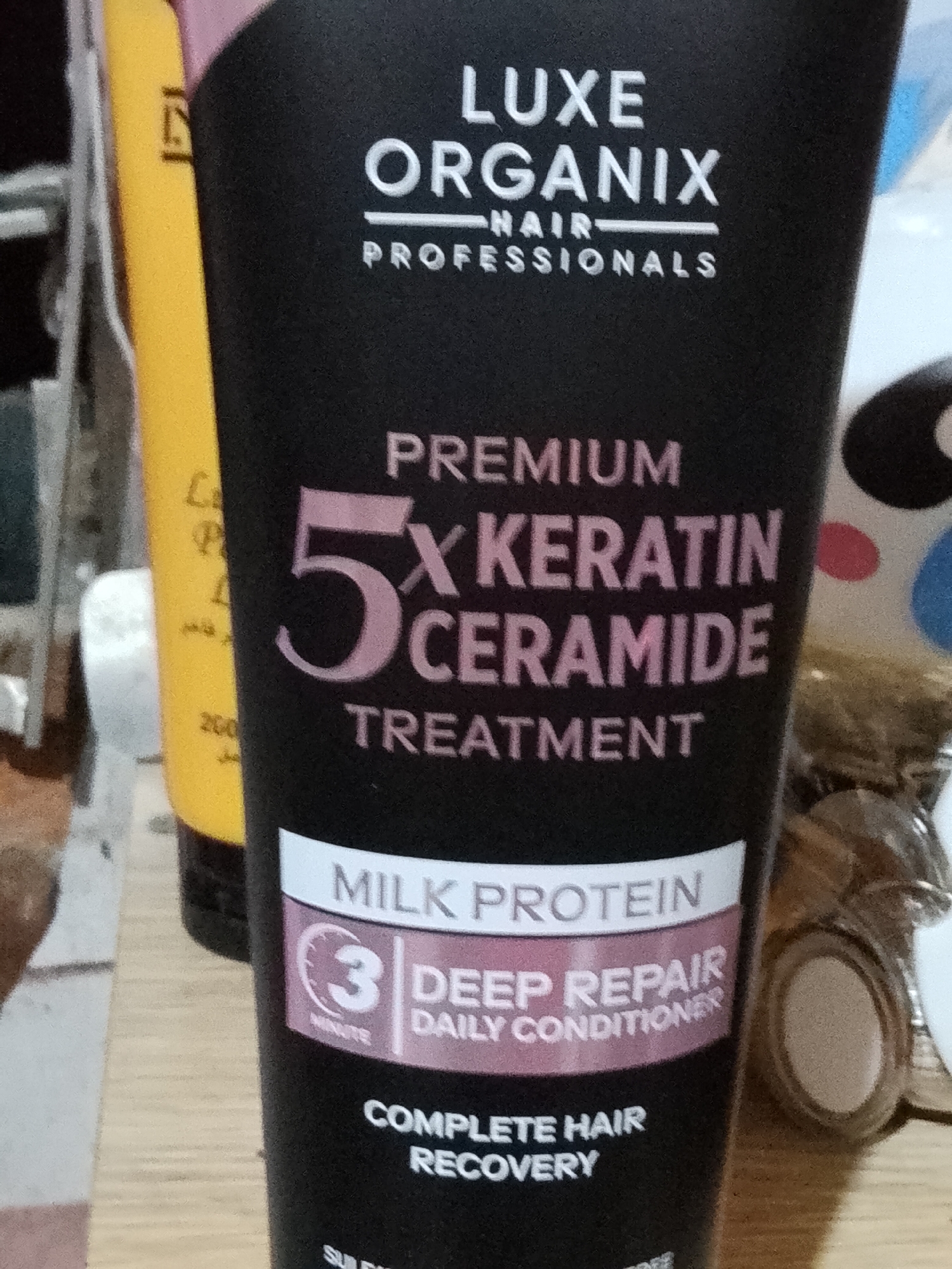 The image shows a close-up of a black hair treatment tube. The tube has white and pink text. The top of the tube reads "LUXE ORGANIX" and below that "NAIR PROFESSIONALS". Below that, it says "PREMIUM 5X KERATIN CERAMIDE TREATMENT". Towards the bottom, it has a pink and white label with "MILK PROTEIN" and a circled "3 MINUTE DEEP REPAIR DAILY CONDITIONER". At the very bottom, it says "COMPLETE HAIR RECOVERY". To the left of the hair treatment tube, a portion of a yellow tube is visible. The background is blurry and appears to be indoors with some colorful circular patterns visible. This image was taken in Davao City, Philippines, on a wooden surface, likely a table, with natural lighting. There are no people visible in the image.