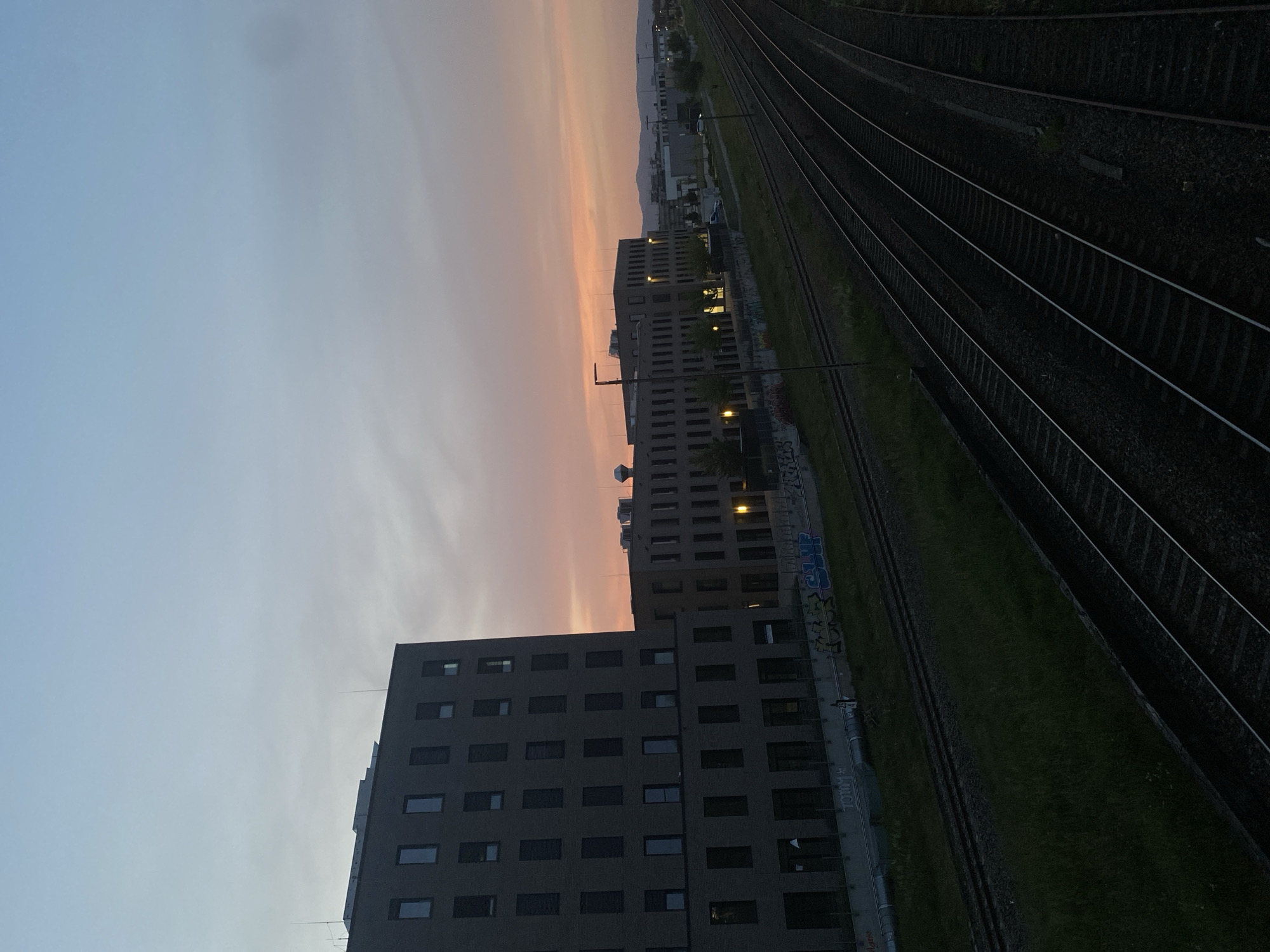 The image shows a view of Landau in der Pfalz, Germany at dusk. Multiple train tracks run diagonally across the right side of the frame, with grass growing between the tracks. To the left, there are several tall, modern buildings with many windows. The sky transitions from blue to orange, indicating a sunset or sunrise. The buildings appear to be dark in contrast to the colorful sky. Graffiti is visible on the side of a building near the train tracks.