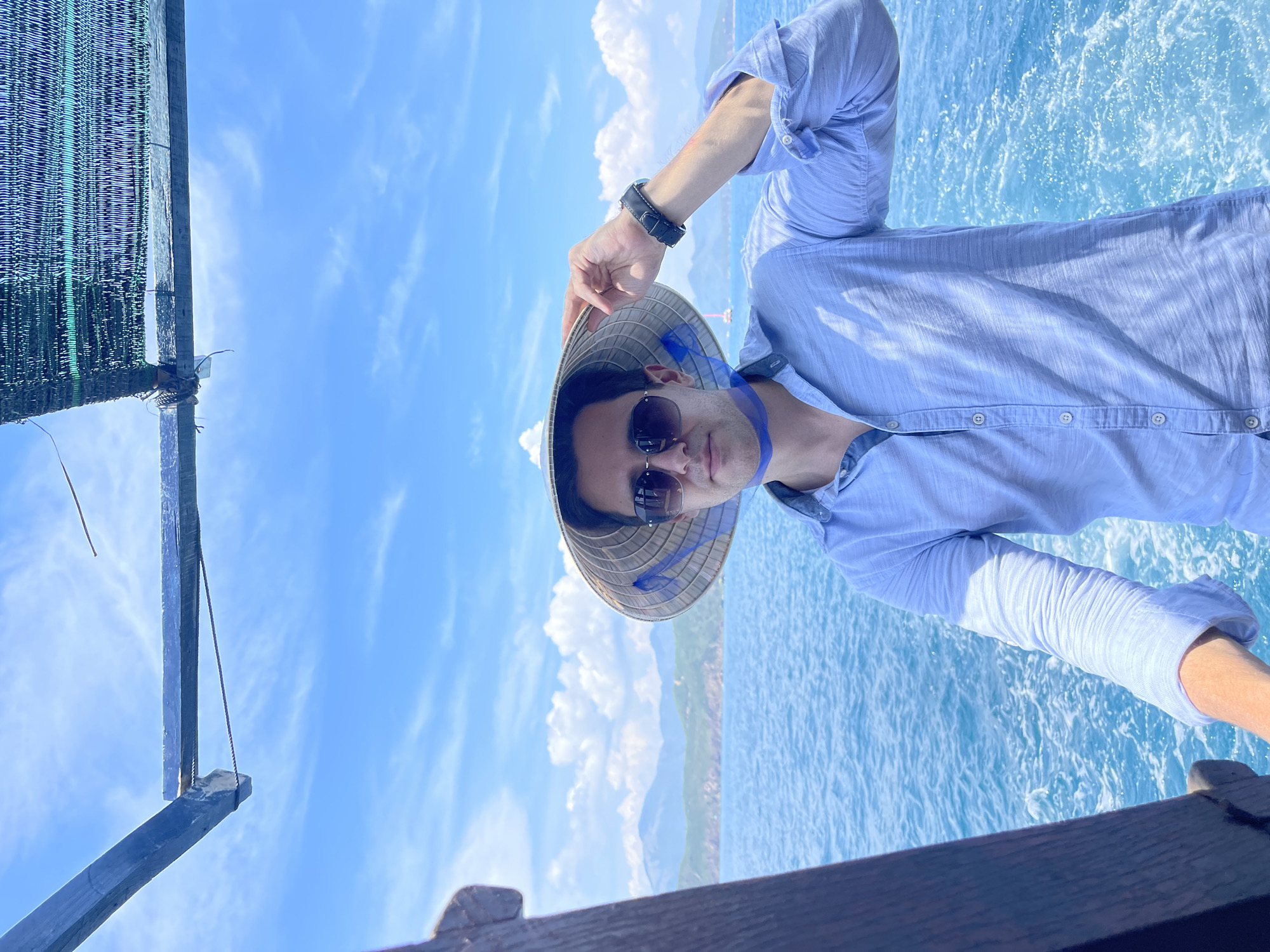 A man wearing sunglasses and a conical hat poses on a boat in Nam Nha Trang Ward, Vietnam. The background shows the blue ocean and cloudy sky. The boat has a wooden railing and a shaded area with netting. The man is wearing a light blue shirt.