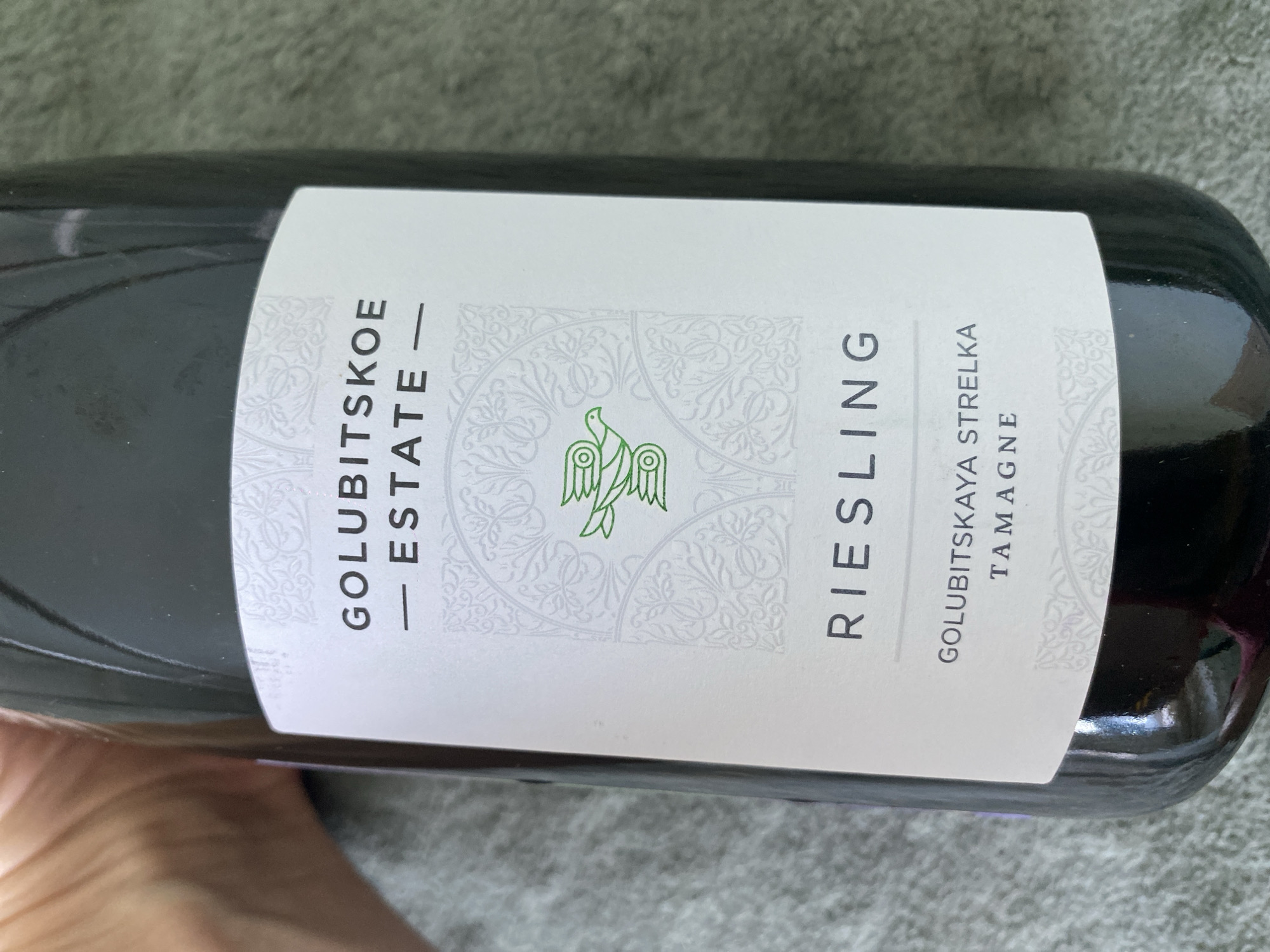 A wine bottle with a light-colored label is held in a person's hand against a textured gray fabric background. The label features the text "GOLUBITSKOE ESTATE" at the top, followed by a green bird graphic with intricate scrollwork. Below the graphic, the word "RIESLING" is printed in large, bold letters. At the bottom of the label, smaller text reads "GOLUBITSKAYA STRELKA TAMAGNE". The bottle is dark glass, and the lighting suggests it is indoors.