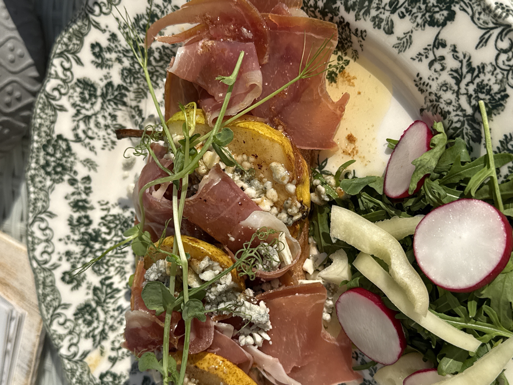 The image shows a plate of food, likely an appetizer or salad, with a focus on the arrangement of ingredients. The plate itself has a white base with a green floral pattern. The food consists of sliced prosciutto, roasted or grilled pear slices filled with blue cheese, pea shoots, sliced radishes, and arugula salad. A light dressing or sauce is visible. The lighting suggests an outdoor or brightly lit indoor environment. There is no indication of the location being Trnava, Slovakia, or any people visible in the image.