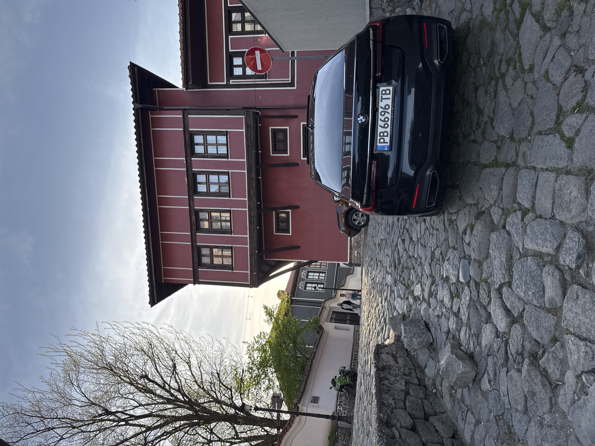 A photograph taken in Plovdiv, Bulgaria, shows a black BMW parked on a cobblestone street next to a red building with white trim, a typical architectural style for the area. A red and white "no entry" sign is attached to the building. Other buildings and trees can be seen in the background. Two people walk along the street further back. A stone wall borders the street, along with a tree in the foreground. The sky is partly cloudy. The license plate on the BMW reads "PB 6696 TB."