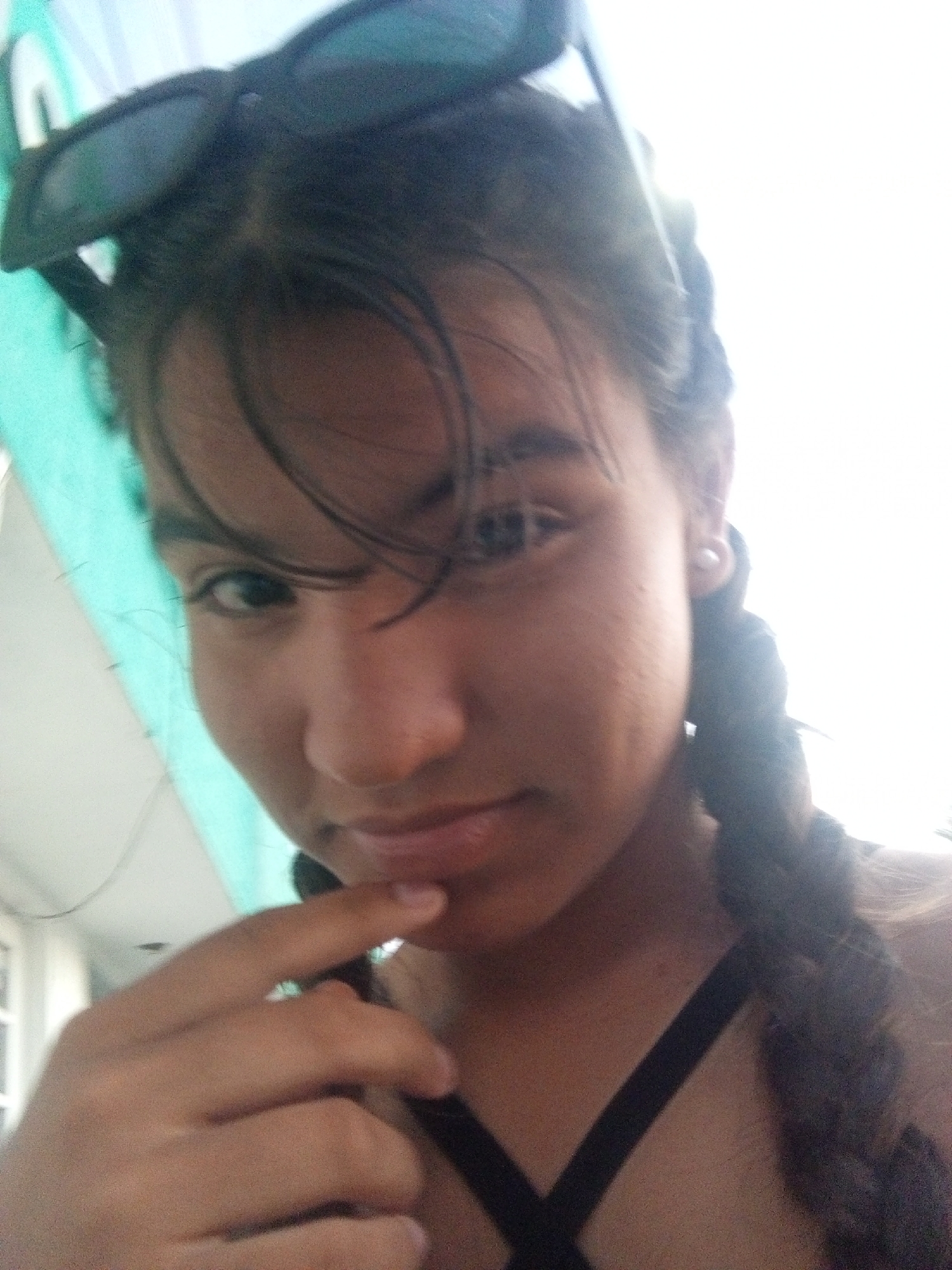 A selfie taken from a low angle features a young woman with dark hair styled in pigtail braids. She has sunglasses perched on top of her head, partially obscuring her forehead. A strand of hair falls across her face. Her right hand is raised, with her index finger touching her chin. She is wearing a black top with crisscrossing straps. A turquoise-colored surface is visible in the upper left corner of the frame. The background is bright and washed out, preventing clear identification of location. It is not possible to confirm the location in Juan Griego, Venezuela, based on visual information.