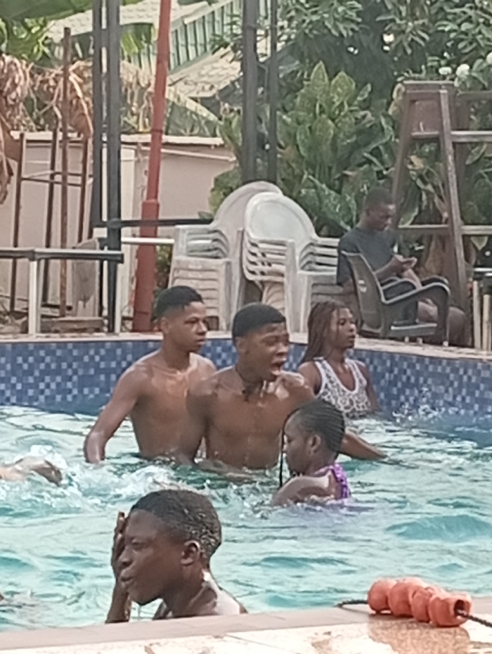 Several individuals are enjoying time in a swimming pool. There are three males, one female, and a young girl. One of the males appears surprised. Another person is sitting on a chair, seemingly occupied with a mobile phone. A ladder and stacked white chairs are visible in the background, along with foliage. Safety floats are positioned at the edge of the pool.
