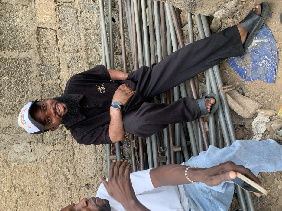 This image captures two adult males outdoors in Okene, Nigeria. The primary subject, positioned right of center, is a young adult male with a dark beard, wearing a white and black baseball cap, a dark ribbed polo shirt with "Reebok" branding, dark trousers, and black slides. He wears a dark digital smartwatch on his left wrist and has his left hand in his pocket. He is seated on a horizontal stack of metal pipes, smiling directly towards the camera. His skin exhibits some lighter discoloration on his forearms.
Partially visible on the left is a second adult male, facing towards the primary subject. He wears a white t-shirt and light blue denim jeans. His right hand is raised and open, gesturing as if in conversation. A light-colored bracelet with multiple small beads is on his right wrist, and he holds a light-colored smartphone in his left hand.
The background consists of a rough, light brown concrete block wall. The men are seated on what appears to be a haphazard stack of metal pipes or scaffolding tubes. The ground beneath them is dirt, littered with small rocks, pieces of broken concrete, crumpled white debris, and a flattened, crumpled blue plastic sheet.