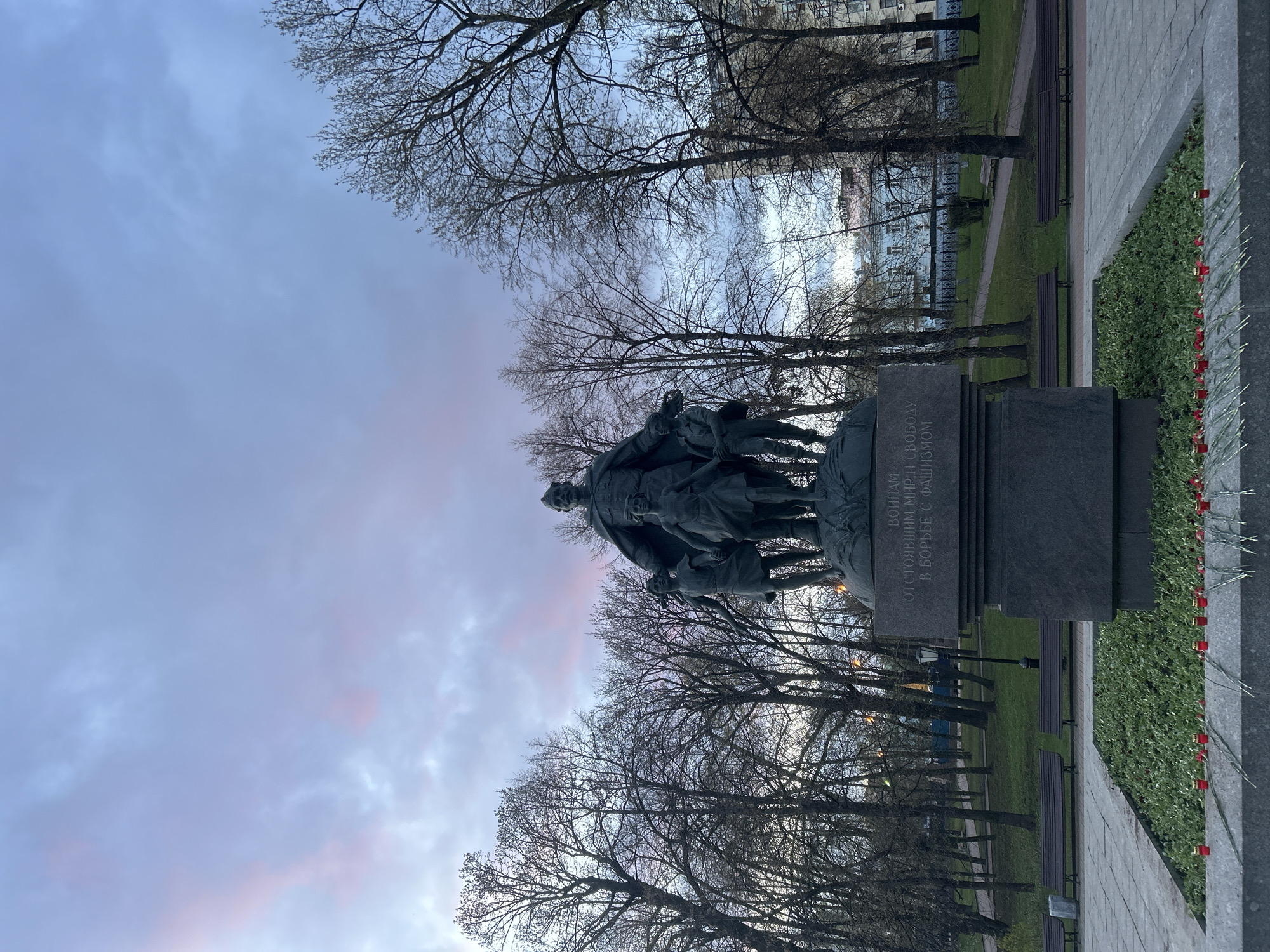 The image depicts a large, dark monument set in an outdoor park or plaza, identified as being in Moscow, Russia. The central subject is a powerful sculptural group, likely made of bronze or dark stone, portraying a commanding male figure, presumably a soldier, standing protectively over smaller figures, possibly children, gathered at his feet. This group rests on a multi-tiered dark stone pedestal.
The surrounding environment consists of a park with numerous tall trees, whose branches are bare, suggesting late winter or early spring. Green grass covers the ground, with paved walkways winding through the area, and several dark park benches are visible. In the background, beyond the trees, light-colored buildings with windows indicate an urban setting.
The sky above is a prominent feature, showcasing soft, diffused light with gentle hues of pink, purple, and pale blue, which might indicate either dawn or dusk, or an overcast day with a specific light quality. The overall lighting is subdued, contributing to a contemplative atmosphere.
A significant detail is a row of small, red objects, resembling candles or possibly carnations, carefully arranged along the edge of a raised flowerbed or wall to the right of the monument, signifying a memorial tribute.
The inscription carved into the side of the monument's pedestal is in Cyrillic. It reads: "ВОИНАМ ОТСТОЯВШИМ МИР И СВОБОДУ В БОРЬБЕ С ФАШИЗМОМ." This translates to "To the Warriors Who defended Peace and Freedom in the Fight Against Fascism," confirming the monument's purpose as a memorial commemorating a historical conflict, most likely World War II. The scene evokes a sense of solemn remembrance and honor for past sacrifices.