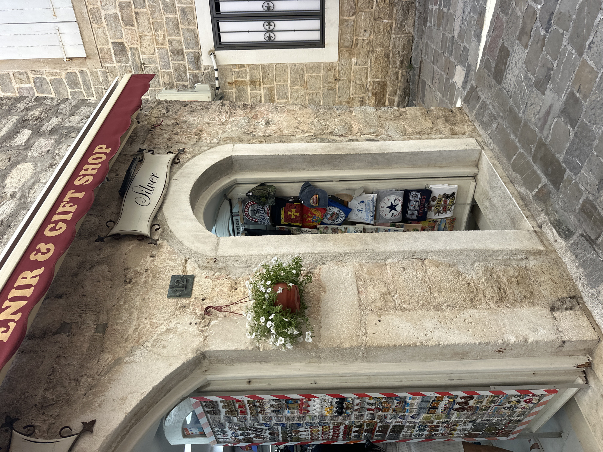 The image captures the facade of a souvenir shop in Budva, Montenegro. The shop's signage reads "SOUVENIR & GIFT SHOP" and "Silver." An arched window displays hats and souvenirs. A hanging flower pot decorates the facade. Another set of souvenirs is displayed beneath the shop's entrance. A building number "12" is visible. The architectural style features stone brickwork.
