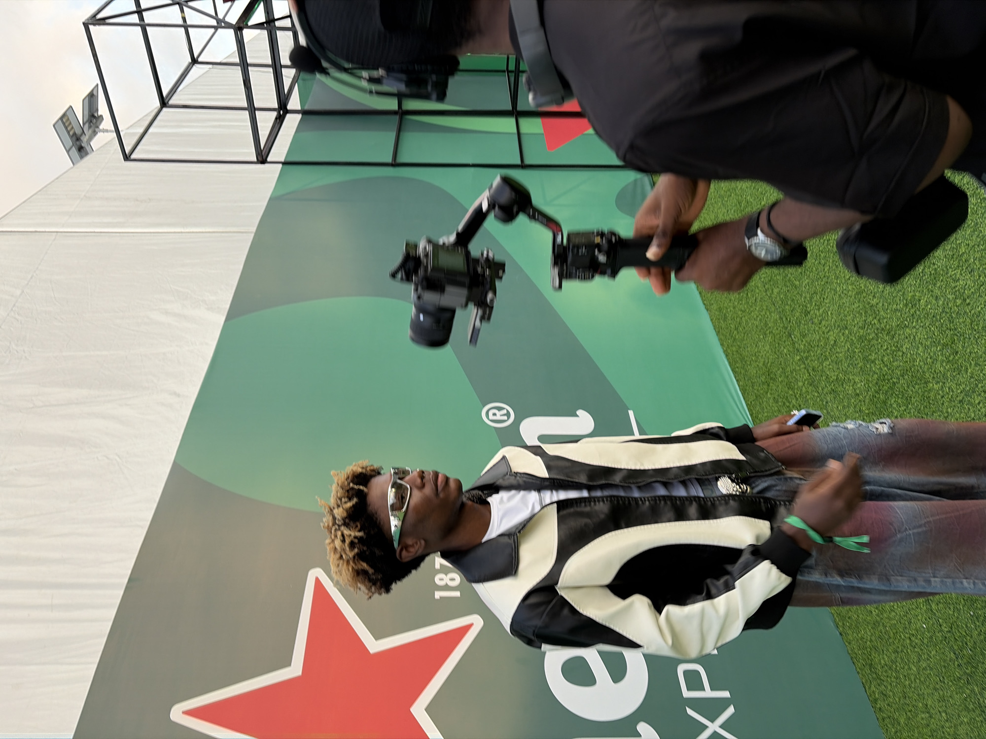 A man with a camera and a gimbal is filming another man with curly blonde hair in front of a Heineken backdrop at an outdoor event. The man being filmed is wearing a white t-shirt, black and white jacket, ripped jeans, and sunglasses. The location is likely Lagos, Nigeria. The environment includes a white tent, green artificial turf, and a metal structure in the background.