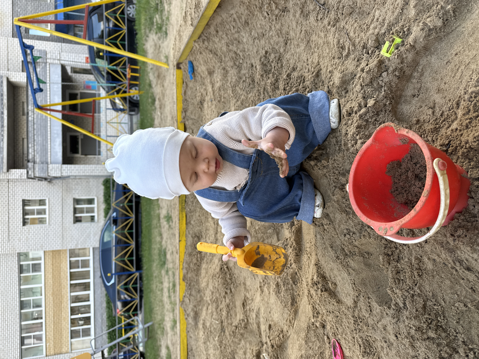 A baby wearing a white hat, a beige sweater, and blue overalls sits in a sandbox, playing with a yellow shovel and a red bucket filled with sand. The baby's eyes are closed, and their right hand is outstretched, holding the shovel. The background shows a playground structure and a building with windows. There are also cars parked near the building. The scene appears to be outdoors, likely during daytime, with natural light illuminating the sand. The ground is covered in sand, with some grass visible at the edges of the sandbox. The overall atmosphere is one of a child enjoying playtime in a playground. The image is oriented vertically, with the top of the frame showing more of the playground and building, and the bottom focusing on the baby and sandbox. There is no visible text in the image.