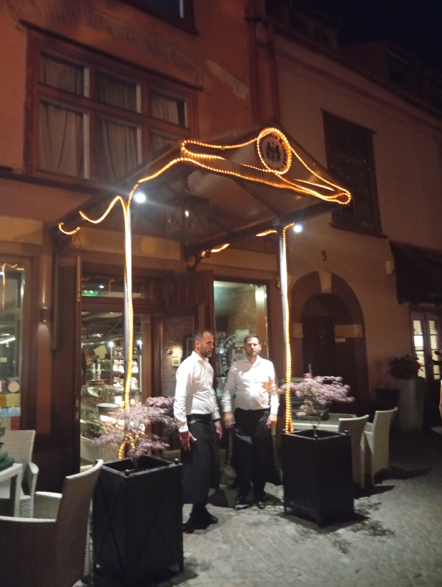 The image depicts the exterior of a restaurant at night, illuminated by decorative string lights. Two servers, dressed in white shirts and black aprons, stand near the entrance. One server is looking away from the camera, while the other faces forward. Outdoor seating with white chairs is visible in the foreground. The building's facade is adorned with ornate windows and a decorative awning. The restaurant appears to be located in the city of Gdańsk, Poland, likely on a cobblestone street, given the context provided.