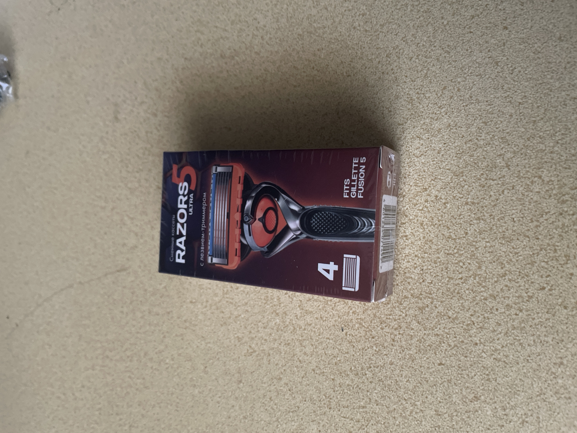 A top-down view shows a box of RAZORS 5 ULTRA razor cartridges on a speckled, beige carpeted surface. The box is dark red and black with a picture of a razor and lettering in Russian and English. Text on the box includes "Сменные кассеты" (Replacement cartridges), "RAZORS 5 ULTRA", "с лезвием-триммером" (with a trimmer blade), "4" with an image of a razor cartridge, and "FITS GILLETTE FUSION 5". The box appears to be sealed with plastic. The carpet has a coarse texture. The lighting suggests an indoor setting. There is no indication of the city of Oryol, Russia, in the image or its context.