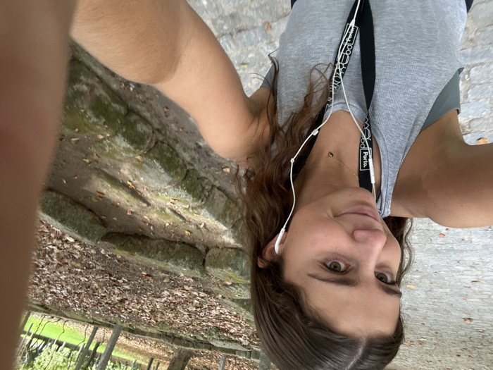 A young female, positioned centrally and angled slightly, captures a close-up selfie in an outdoor park in Aldoar, Foz do Douro e Nevogilde, Portugal. She has medium-brown wavy hair, dark eyes, and a subtle smile, looking directly at the camera. She wears a grey ribbed tank top over a darker undergarment, with white wired earphones inserted into both ears. A black lanyard featuring "Porto." text and a geometric pattern in white is visible around her neck, entwined with the earphone cable. A delicate gold-colored necklace with a small dark spherical pendant is also worn. Her right arm is extended, holding the camera, with portions of her left hand visible at the bottom right. The immediate foreground shows a grey cobblestone path. The background on the right reveals a dirt embankment featuring several layers of moss-covered, rough-hewn stone blocks. The ground of the embankment and portions of the path are covered with scattered dry brown leaves. Trees with green and sparse foliage are visible further back, beyond which a patch of green grass indicates an open area. The scene is illuminated by natural daylight.