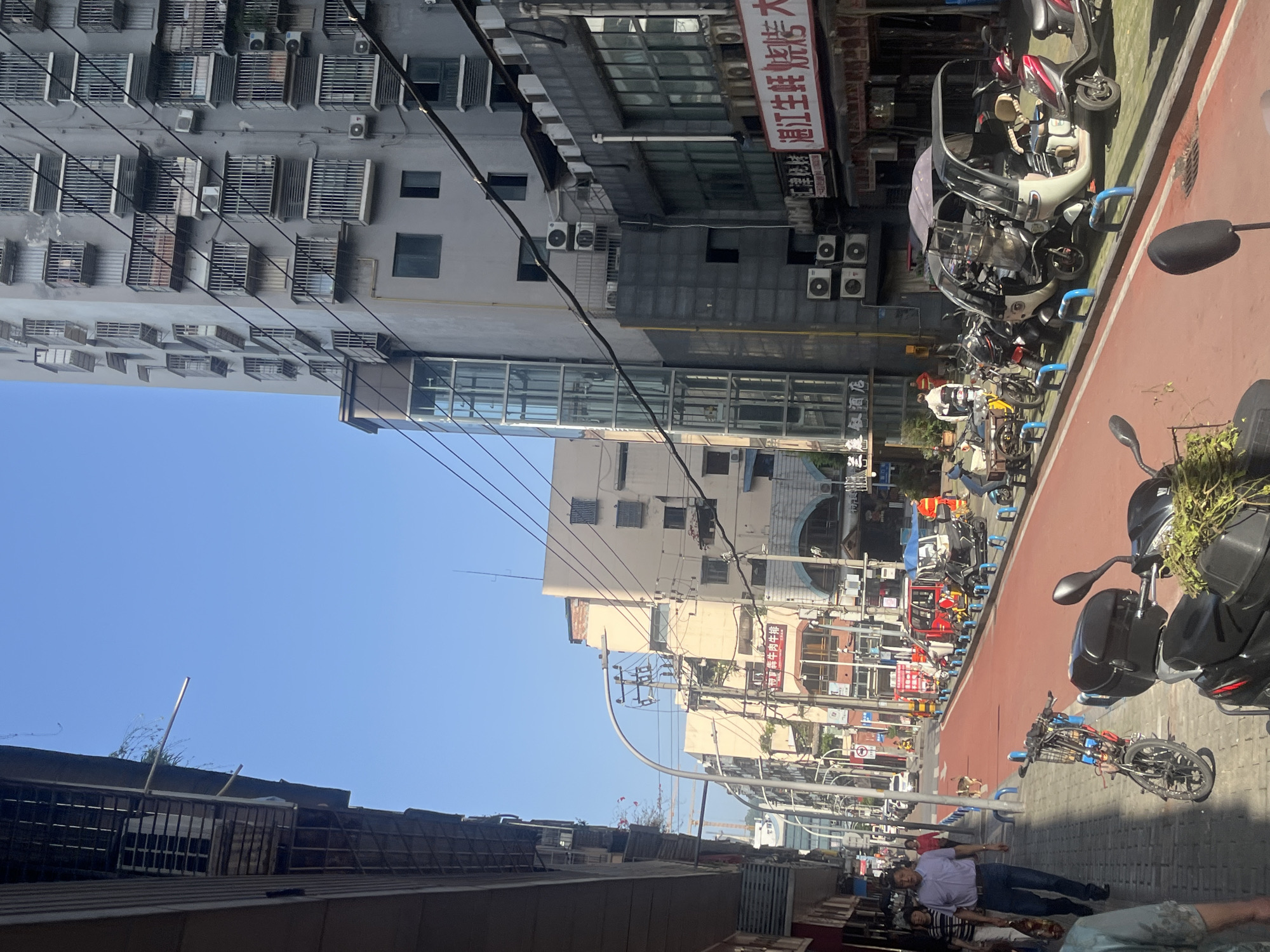 This photograph captures a vibrant street scene in an urban environment, likely in Jiangjin, China, given the provided context. The image is oriented with the clear blue sky on the left, but if rotated clockwise, it depicts a street stretching into the distance under a bright, sunny sky.
On the right side of the street, a distinct red paved path, possibly a dedicated lane for bicycles or scooters, is visible. Adjacent to it, a grey sidewalk is lined with numerous parked electric scooters and motorbikes, some featuring protective canopies and blue U-shaped bike racks. Further down, a blue bicycle is parked. A man in a light purple shirt and dark pants is standing on the sidewalk in the foreground, with another person partially visible beside him.
Buildings of varying heights and styles line both sides of the street. On the left, a tall, multi-story residential or commercial building dominates, featuring numerous windows, many with metal security grates, and a multitude of outdoor air conditioning units. Utility poles laden with intricate networks of overhead wires are prominent, crisscrossing the frame. On the right, further down the street, other buildings with various signs and architectural details can be seen.
Visible text on a red banner affixed to one of the buildings reads "湘江生鲜烤肉大" (Xiangjiang Fresh BBQ Restaurant). A smaller sign below it appears to say "酒店" (Hotel). The scene suggests a bustling urban area during daylight hours, with no specific event unfolding beyond routine daily life. The clear sky indicates good weather.