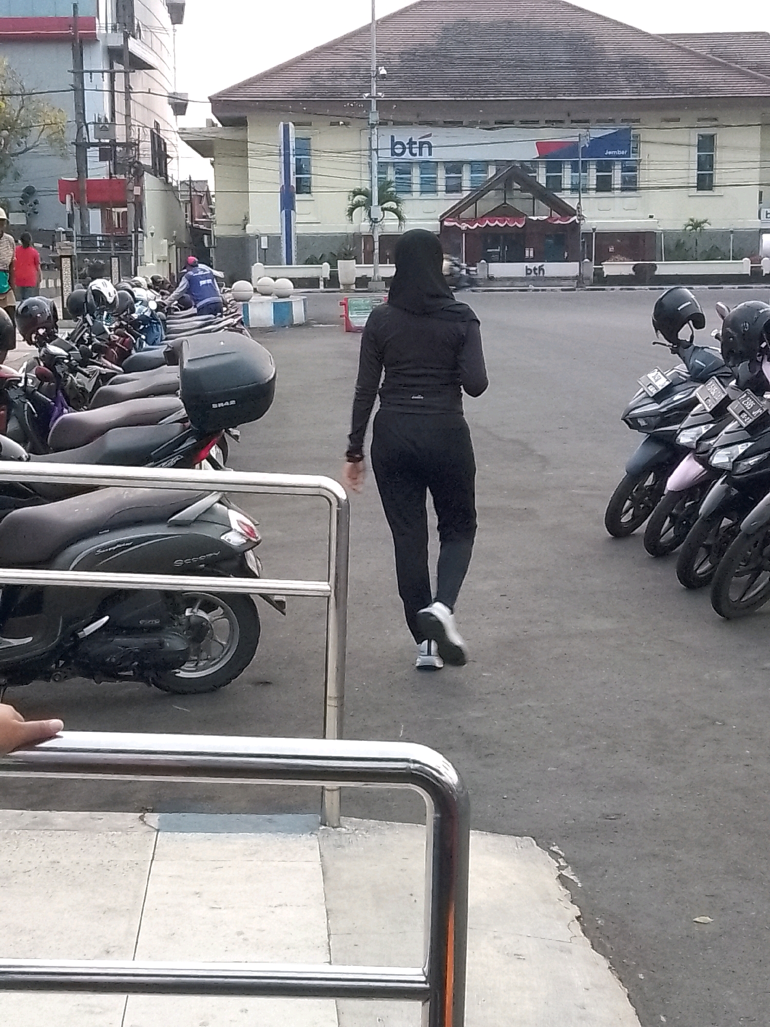 In Jember, Indonesia, a woman wearing a black hijab and athletic clothing walks away from the camera on a paved surface. She is wearing dark pants and a black long-sleeved top, with white and grey athletic shoes. In the background, a line of parked motorcycles is visible, with a building behind them displaying the "BTN" logo and "Jember" text. The building appears to be a financial institution. The overall scene suggests an urban environment in Jember.