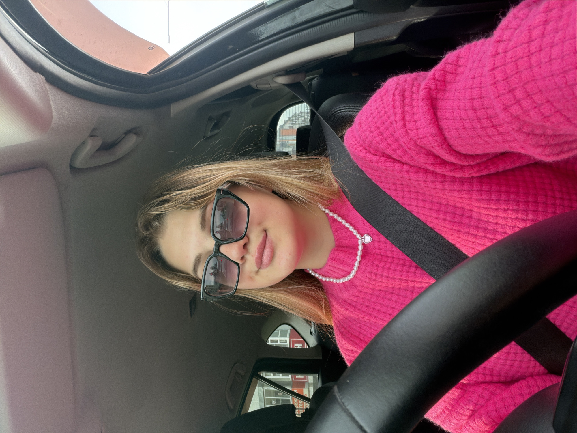 A young woman wearing a bright pink sweater, a pearl necklace with a heart pendant, and sunglasses is sitting in the driver's seat of a car. She is wearing a seatbelt. The interior of the car is visible, including parts of the steering wheel, dashboard, and ceiling. Outside the car windows, buildings and street scenes are visible, which, based on the presence of Cyrillic script and the style of architecture, indicate a location in Agryz, Russia. The image appears to be taken from the perspective of the driver, and it may be a selfie.