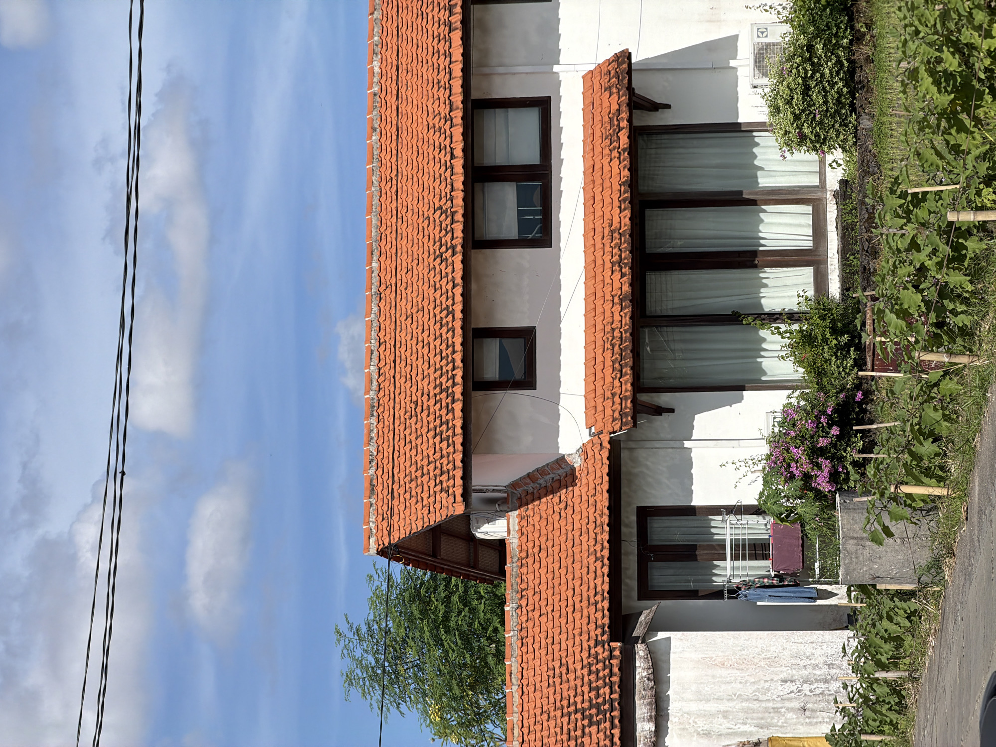 The image shows a building with a red-tiled roof in North Kuta, Indonesia. The building is white with brown-framed windows and an air conditioning unit is mounted near the top. Lush greenery including various plants and flowers surrounds the base of the building. Laundry hangs on a drying rack outside one of the windows. Electrical wires stretch across the blue sky with scattered clouds.
