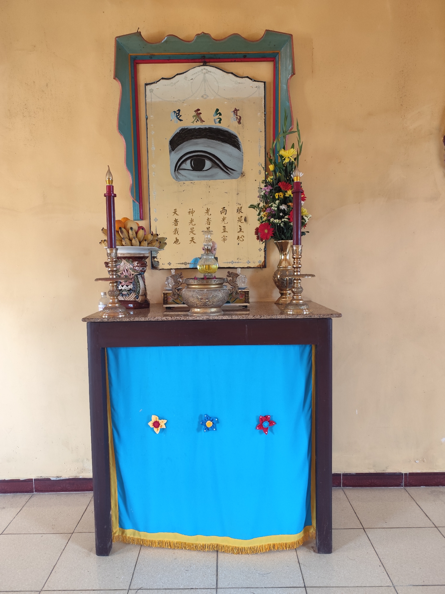 A table with a marble top is set up against a pale yellow wall. On the table, an altar is arranged with religious items. A framed painting of an eye with Chinese calligraphy is mounted on the wall above the altar. To the left of the painting is a red candle in a decorative holder, and a bowl of bananas. To the right of the painting, a vase holds a bouquet of colorful flowers. In front of the altar, the table is draped with a bright blue cloth adorned with three felt flowers: a yellow and red one on the left, a blue one in the middle, and a red one on the right. The cloth is trimmed with yellow fringe at the bottom. The floor is tiled with light-colored square tiles. The scene suggests a religious or spiritual space, possibly in the city of Lộc Quý, Vietnam, given the mention of the location.