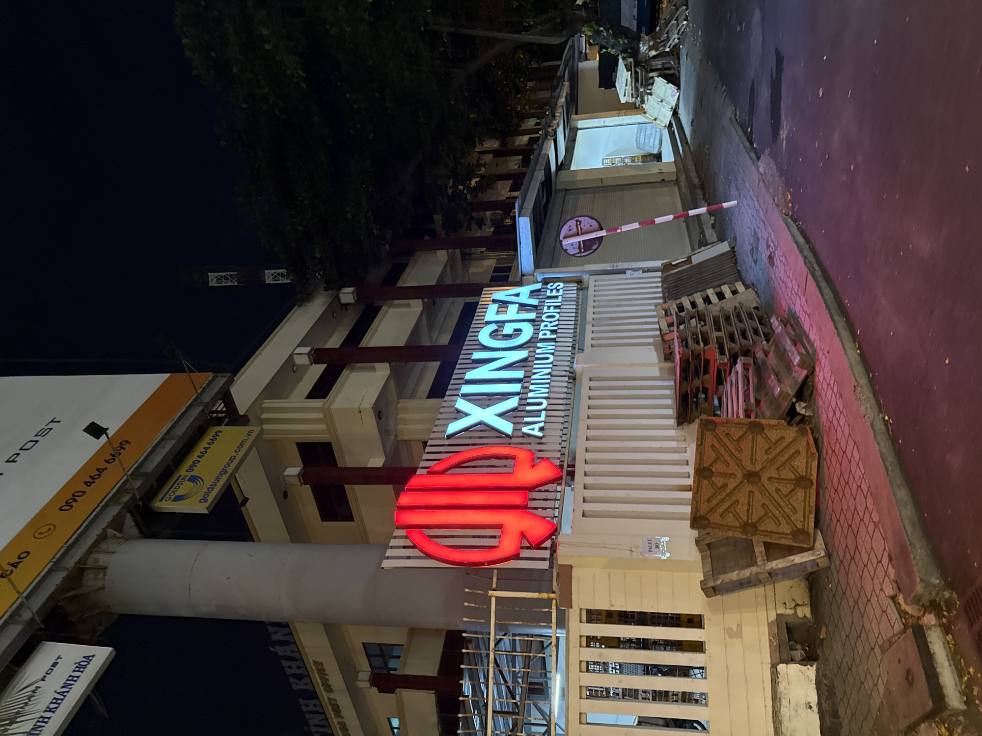The image depicts a nighttime scene in Nha Trang, Vietnam, focusing on the exterior of a building with a prominent illuminated sign. The sign, glowing in a vibrant red and bright cyan, reads "XINGFA ALUMINIUM PROFILES" and is mounted on a white, slatted fence. To the left of the sign, a large red abstract logo is visible. Above the fence, a section of a building with pillars and windows is seen.
In the foreground, a sidewalk is paved with reddish-brown bricks, partially covered by fallen leaves. Scattered on the sidewalk and against the building are several wooden pallets, some stacked and others lying flat. One pallet, in the center, is particularly visible and appears to be made of compressed wood fiber. A metal pole with alternating red and white stripes stands vertically near the pallets.
To the right of the building entrance, a roll-up door is visible, partially closed. Beyond the door, the interior of the building is illuminated. Further to the right, stacks of wooden pallets are visible, along with a dark-colored vehicle.
Above the building, a large billboard is partially visible, with text in Vietnamese and English, including a phone number. A streetlamp casts light on the scene. The overall environment suggests an industrial or commercial area at night.
