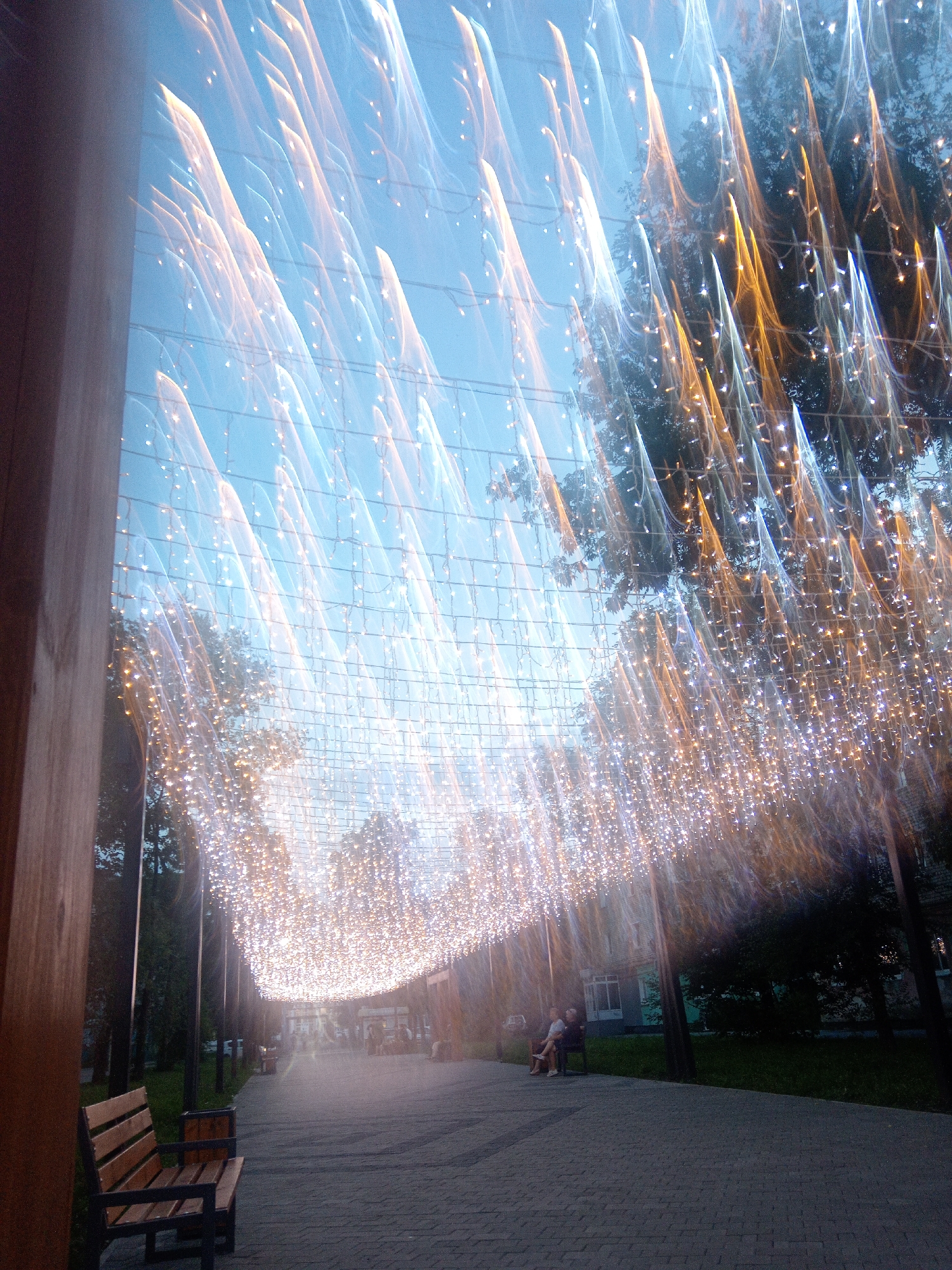 The image depicts an outdoor pedestrian walkway illuminated by strings of lights creating a canopy effect, likely in Belogorsk, Russia. The lights are blurred due to motion or long exposure, giving a streaky appearance. Trees line the walkway, and a paved path is visible, with a wooden bench in the foreground on the left. In the middle ground, two individuals are seated on a bench on the right side of the walkway. Buildings and more trees are visible in the background. The overall atmosphere is one of an evening or twilight setting with festive lighting.