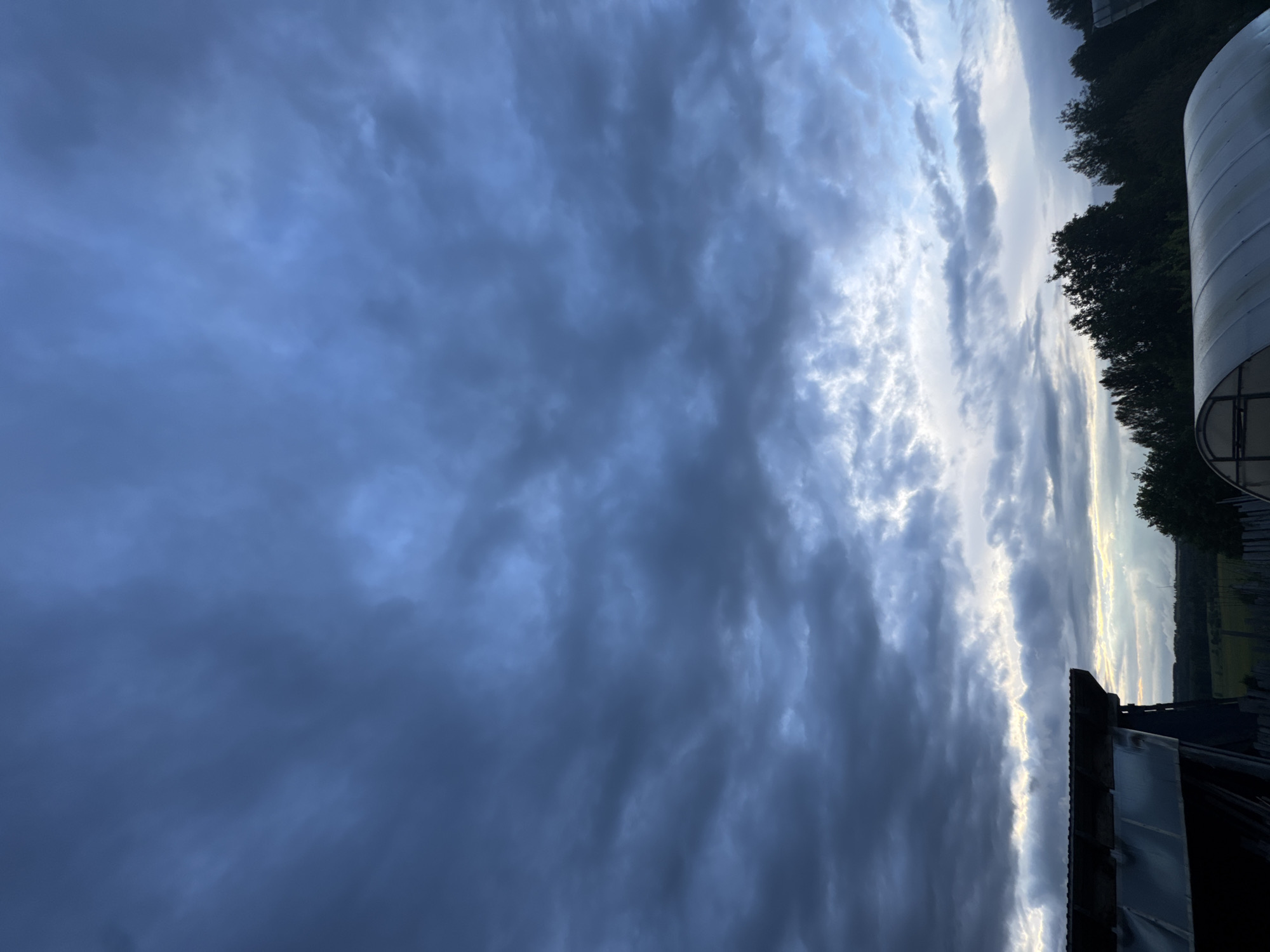 The image shows a cloudy sky over Nizhnyaya Gar', Russia. The sky is predominantly filled with dark, grey clouds on the left side. A bright, narrow opening of lighter, white clouds cuts through the center of the image. The right edge reveals dark green trees and a section of a white, cylindrical structure. A small portion of a roofline is visible in the lower right corner.