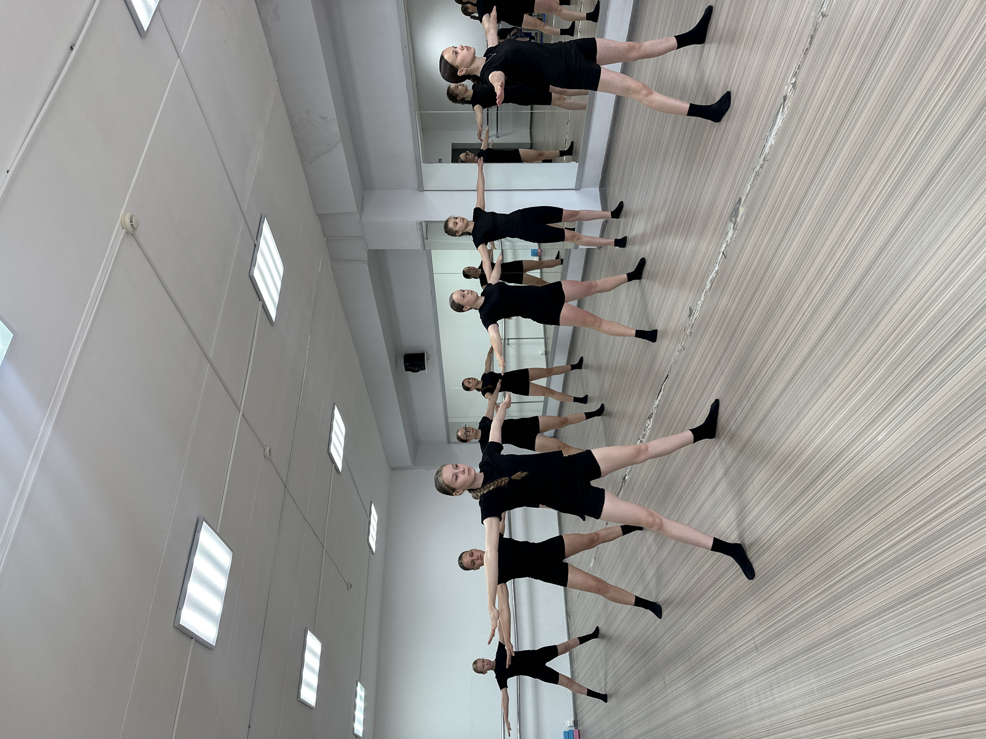 In a dance studio located in Votkinsk, Russia, a group of seven young girls, appearing to be between the ages of 8 and 15, are engaged in a dance exercise. They are all dressed in matching black leotards and shorts, with black socks. The studio has a light-colored, wood-paneled floor and white walls. Fluorescent ceiling lights illuminate the space. Large mirrors line one wall of the studio, reflecting the dancers and the room. The girls are positioned in a staggered formation, facing the viewer, with their arms extended horizontally to the sides, palms down, and their legs spread in a wide stance. Their expressions are focused and intent. The overall impression is one of a structured dance class.