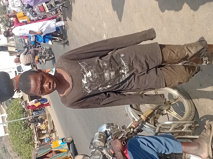 A young man in a dark long-sleeved shirt and pants is standing in front of a parked motorcycle. He appears to be on a street, with other motorcycles and people visible in the background. The scene is outdoors on a sunny day. He is wearing sandals and looking at the camera. Buildings and vehicles are seen blurred in the distance.