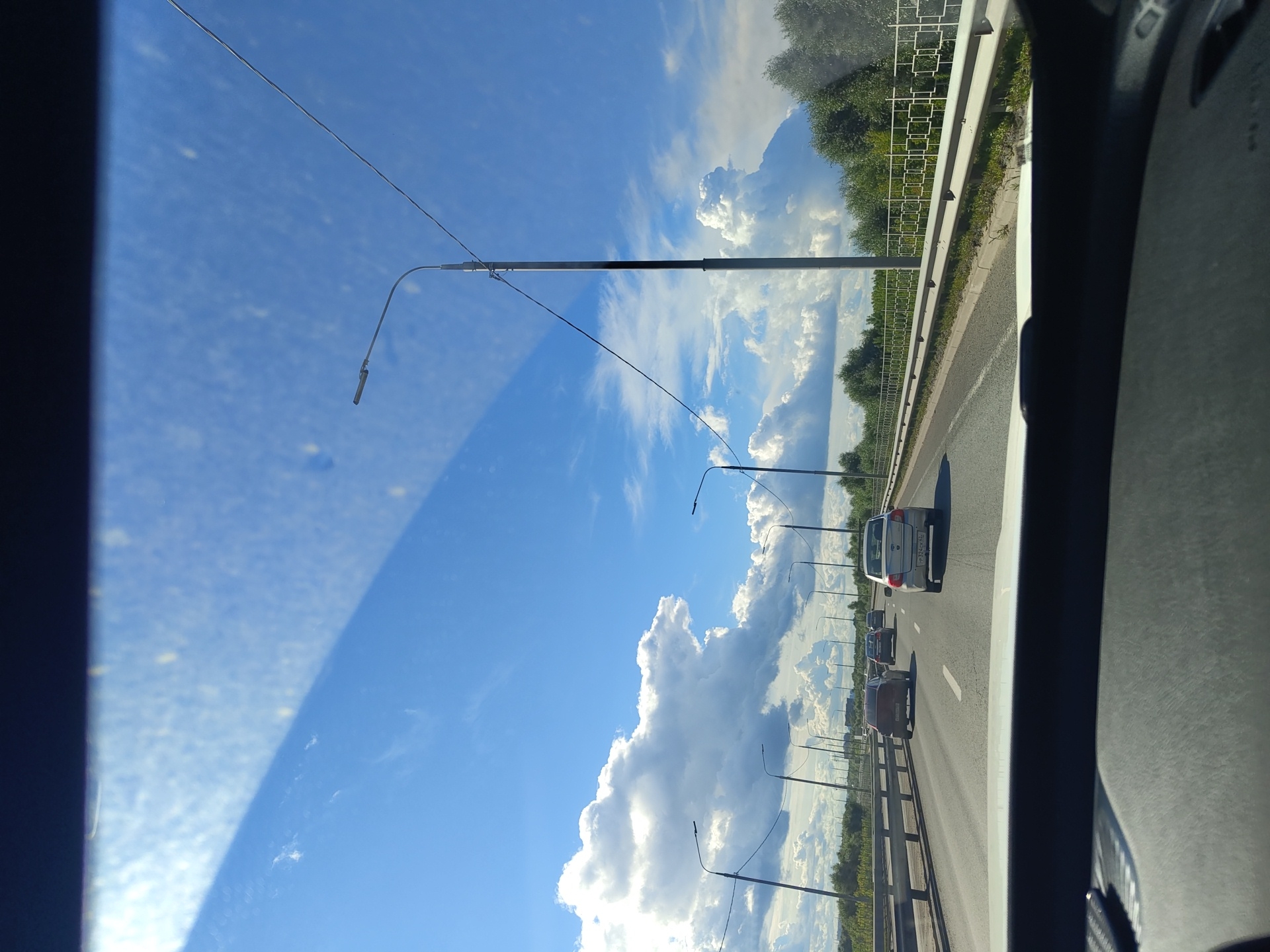 The image captures a view from inside a vehicle looking out through the windshield at a highway scene in Фруктовая, Russia. The sky is predominantly blue with large, puffy white clouds. Several streetlights line the right side of the highway, and a power line is visible running diagonally across the sky. Trees and foliage form a green border along the right side of the road. Multiple cars are driving in the same direction on the highway, with traffic appearing moderate. The dashboard of the vehicle is visible at the bottom of the frame. The overall impression is a sunny day on a suburban or rural highway.