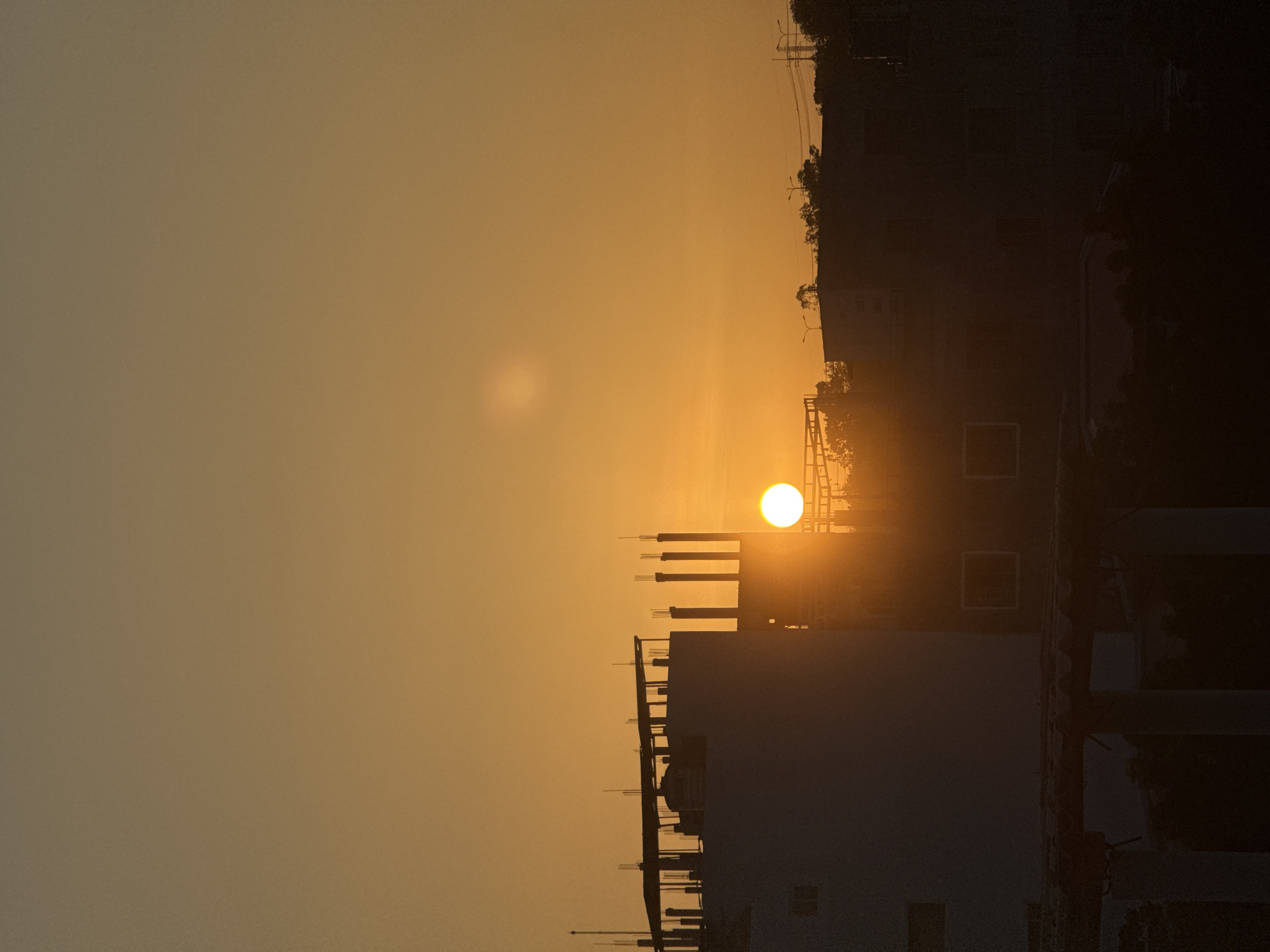 A vertical, medium shot captures the sun setting behind buildings in Mũi Né, Vietnam. The sky is a gradient of warm orange hues, with a brighter, slightly hazy sun appearing as a perfect circle on the right side of the frame. In the foreground, silhouetted against the sunset, are several buildings. On the left, a white building with visible rebar and scaffolding on its rooftop dominates the frame. To its right, another building is visible, also in silhouette, with multiple vertical pillars extending from its roof. The lower portion of the image is filled with the dark silhouettes of more structures and possibly foliage, obscured by the low light. No people or specific actions are discernible due to the silhouette effect and the time of day. The scene suggests an urban or semi-urban environment at dusk.