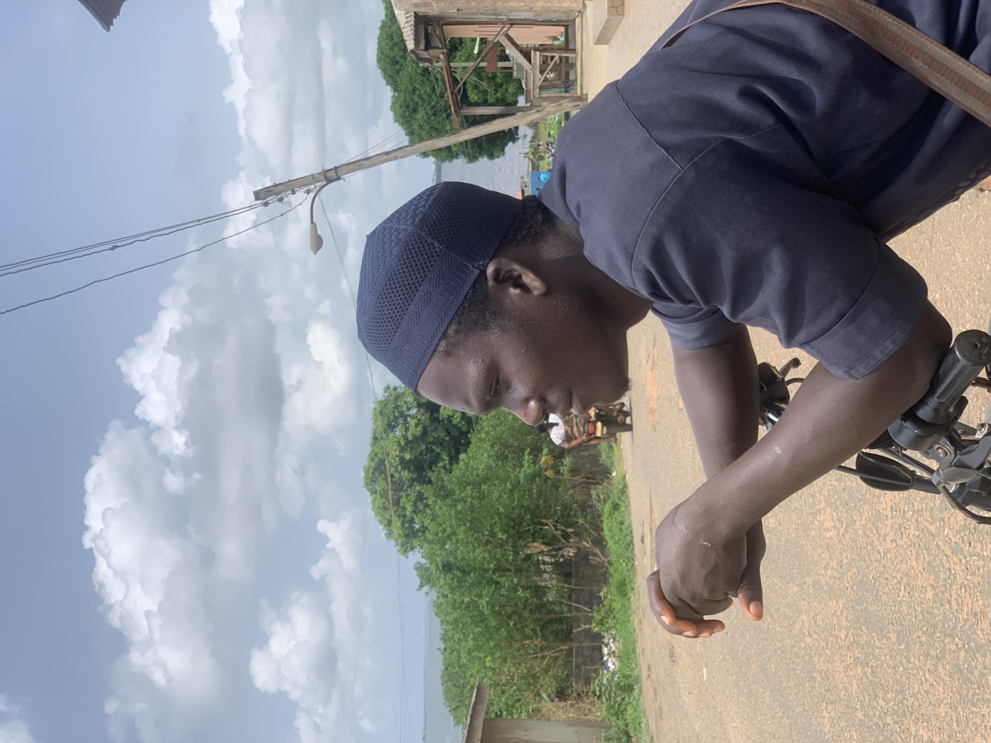 A person wearing a dark blue kufi cap and matching short-sleeved shirt is leaning over the handlebars of a motorcycle. The individual's hands are clasped together on the handlebars. In the background, there is a body of water partially obscured by trees and vegetation. A utility pole with electrical wires and a streetlight is visible on the left side of the frame. A small, wooden structure is visible near the body of water. The sky is overcast with numerous clouds. The location appears to be a street in Ikorodu, Nigeria.