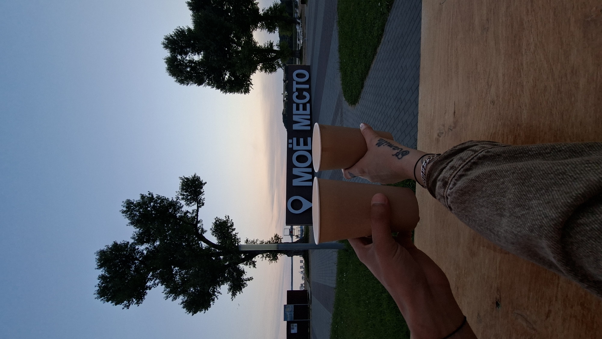 Two people's hands hold paper cups in the foreground of the image. The cup in the left hand is larger and appears to be held by a male based on the visible arm and skin tone. The cup in the right hand is smaller and held by a female, as indicated by the visible arm, skin tone, and a tattoo that reads "Kamilla" on her inner wrist, along with a bracelet. The background features two trees on either side of a large sign that reads "МОЁ МЕСТО" in Russian, which translates to "MY PLACE". The location is identified as being in the city of Myadzyel, Belarus. The sky is a dusky blue, indicating either dawn or dusk, with a hint of pink and orange on the horizon. Beyond the sign, a body of water and beach chairs are visible, suggesting a waterfront location. The ground in the foreground is a wooden surface.