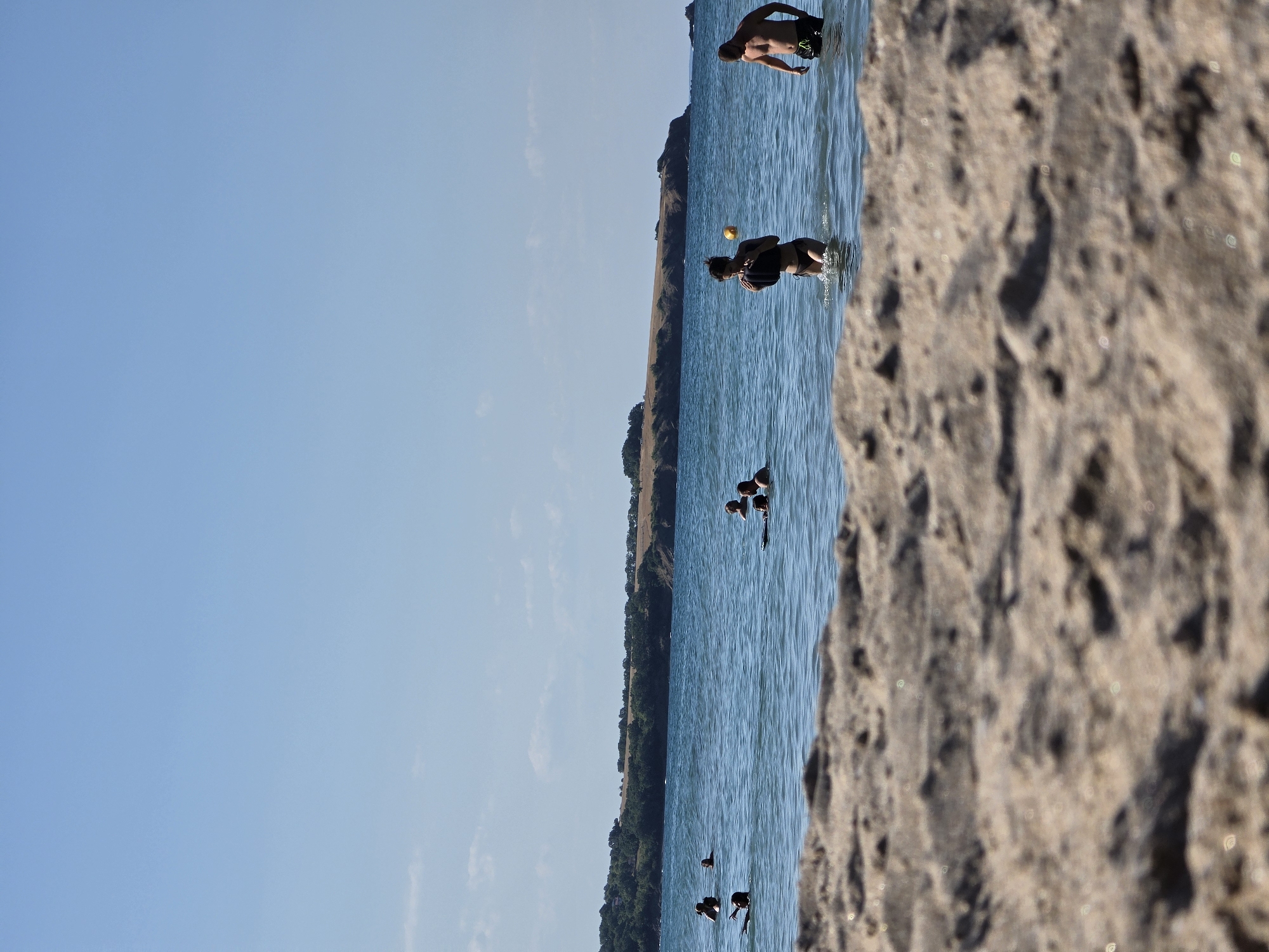 The image depicts a beach scene in Ahtopol, Bulgaria. In the foreground, the sandy beach is blurred and out of focus. The midground shows a body of water with several individuals swimming and wading. On the right side of the water, a man in dark swim trunks stands partially submerged, facing left. To his left, a woman in a black bikini is wading out of the water, holding a black object to her chest, with a yellow ball visible in the water to her right. Further out in the water, other people are visible, some swimming and one person on a surfboard. In the background, a long, treed coastline stretches across the horizon under a clear blue sky with a few scattered clouds. The sea is calm with gentle waves.