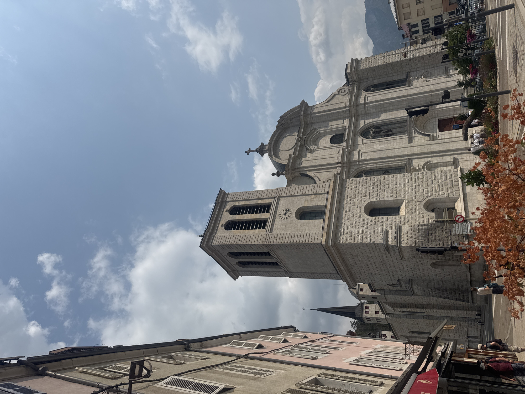 In Saint-Claude, France, an exterior shot captures a view of the Saint-Pierre Cathedral. The cathedral, constructed of light-colored stone, features a tall bell tower with arched openings and a clock face, along with a larger, more ornate main structure topped with a cross. To the left of the cathedral, a street is lined with buildings, one painted pink and another a muted gray, featuring windows with shutters. Several individuals are visible on the sidewalk, some interacting near shop entrances. Further down the street, traffic lights and street signs are present. The scene is set against a bright, cloudy blue sky, with distant mountains visible in the background.
