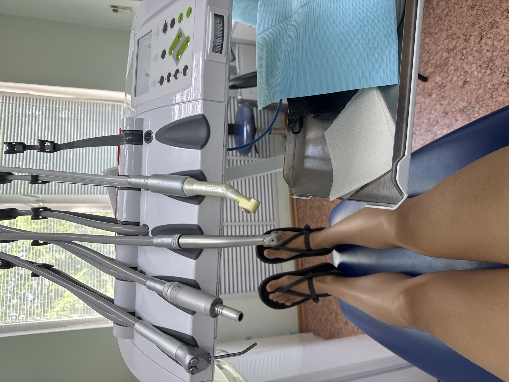 The image shows a person sitting in a dental chair, viewed from above. The person is wearing flip-flops and has their legs extended. In front of them is a dental instrument panel with various tools and controls. The panel is white with gray accents and has several attached instruments. The chair is blue and appears to be in a dental office setting.