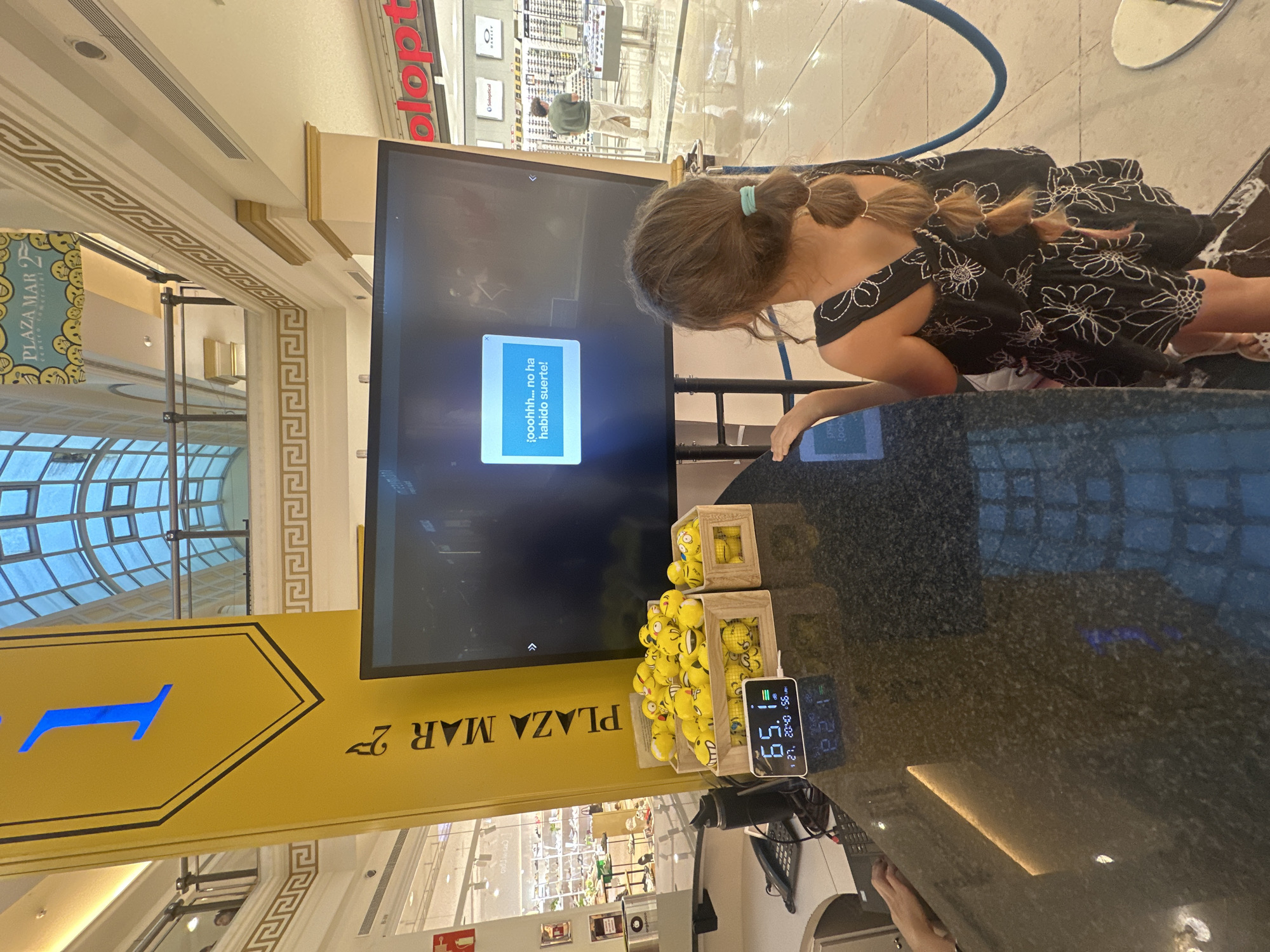The image, taken in what appears to be a brightly lit shopping mall or commercial center, shows a young girl facing a large digital screen. The perspective is rotated, with the floor and ceiling appearing on the sides.
The girl, with her back to the viewer, has light brown hair tied in two pigtails, one secured with a light blue elastic. She is wearing a dark-colored dress or top with white floral patterns. She is leaning on a dark, speckled countertop or desk, with her right arm visible.
The central object is a large, dark-screened monitor, possibly an interactive display. On its screen, a blue pop-up box reads "¡ooohhh... no ha habido suerte!" which translates to "Ohh... no luck!". This suggests the girl might have been participating in a game or a drawing.
On the counter next to the screen, there are two wooden boxes filled with numerous small, round, yellow plush toys resembling emoji faces, mostly smiling or laughing. To their right, a digital display shows "65.1", "21, 20:40 56", and "22:1", likely indicating temperature, date, and time (8:40 PM on the 21st). A coiled light blue cable is also visible.
The setting is characterized by elaborate architecture, including a domed skylight with a grid pattern and decorative friezes with Greek key patterns along the walls and ceilings. A yellow sign points left with "PLAZA MAR 2" visible in reverse. Another sign in the background reads "PLAZA MAR" and part of "plopt", possibly an optical store name, with "Soloptical" and "OAKLEY" branding visible in the distance. The flooring is a light-colored tile, reflecting the overhead lights. The overall impression is an evening scene within a modern commercial complex.