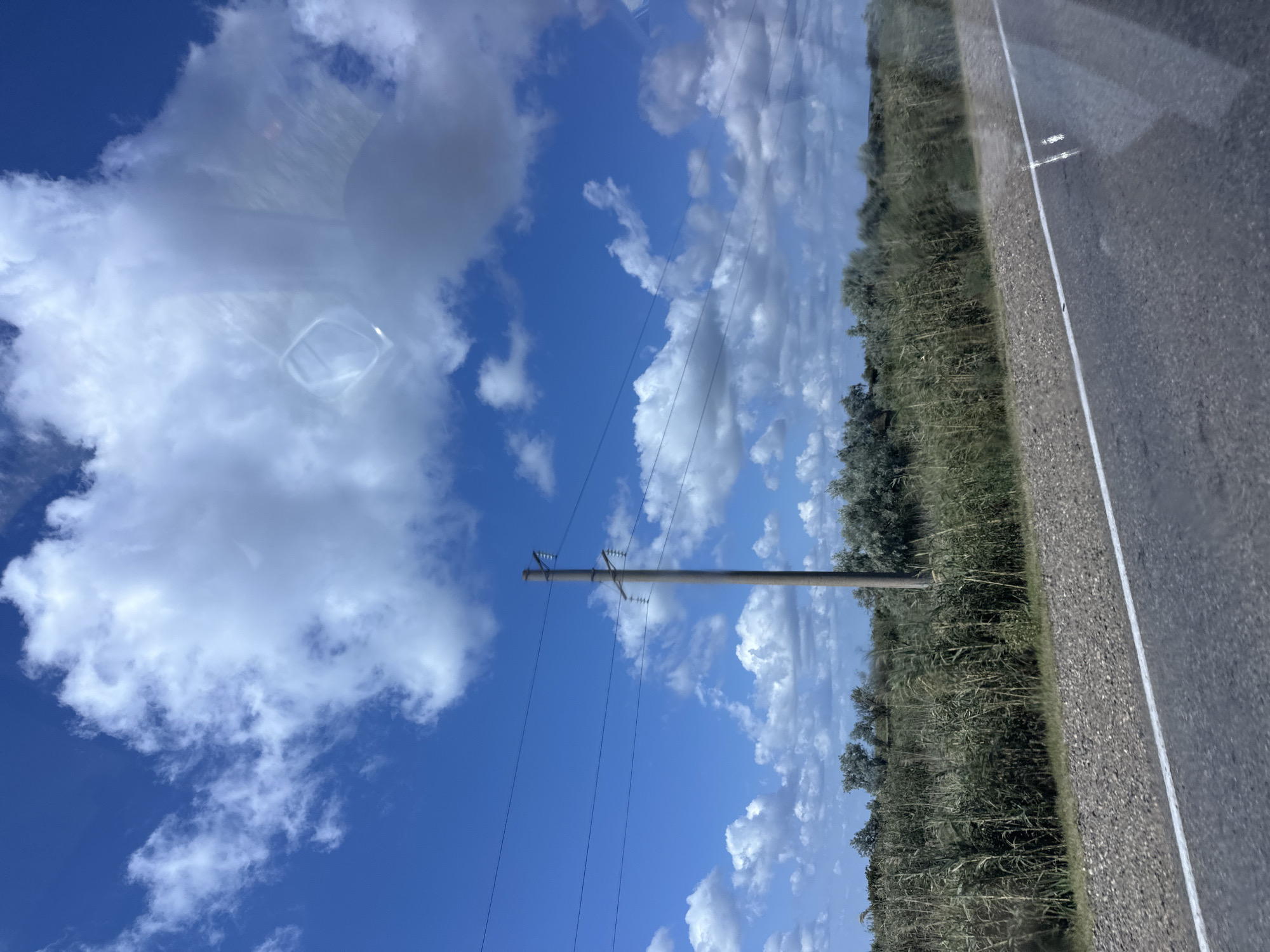 The image shows a roadside view with a clear blue sky filled with white, fluffy clouds. A single utility pole stands on the right side of the frame, supporting several power lines that stretch across the sky. In the foreground, a paved road with a white line marking the edge is visible, bordered by a gravel shoulder. Beyond the shoulder, there is a dense line of green vegetation, appearing to be tall grass or reeds, with some smaller trees in the distance. The overall scene is outdoors, under a bright sky, likely in a rural or semi-rural area, as suggested by the vegetation and utility pole. The location in Peresyp, Russia, suggests a coastal or steppe environment, consistent with the vegetation and open sky. There are no people or animals visible in the image. The image appears to have been taken from a moving vehicle, as indicated by the slight blur and reflections on the right side of the frame.