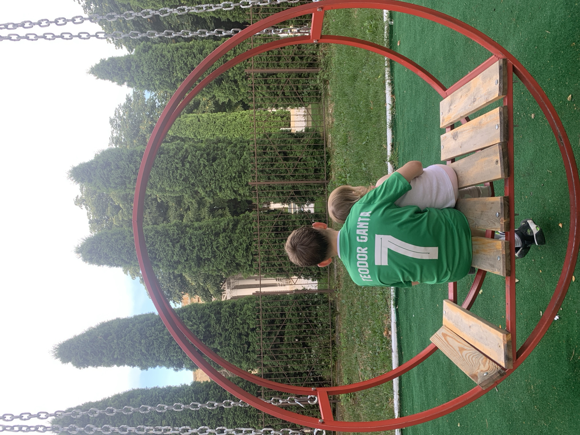 This media file shows a view from behind of two children sitting on a wooden bench attached to a red, circular swing frame. The older child, a boy wearing a green soccer jersey with the name "TEODOR GENTA" and the number "7" visible on the back, has his arm around the younger child, a girl. They are seated on the bench, facing away from the camera, with their backs to a metal fence and lush green trees and bushes. The ground is covered with green artificial turf, with patches of natural grass visible in some areas. A white line is also visible on the turf, suggesting a sports field. The sky appears bright, indicating it is daytime, and the weather seems pleasant. The setting appears to be a playground or park in Dondușeni, Moldova, given the general appearance of the trees and the presence of what looks like a building behind the fence. The posture of the children and their close proximity suggest a moment of companionship and comfort, possibly after playing or while resting.
