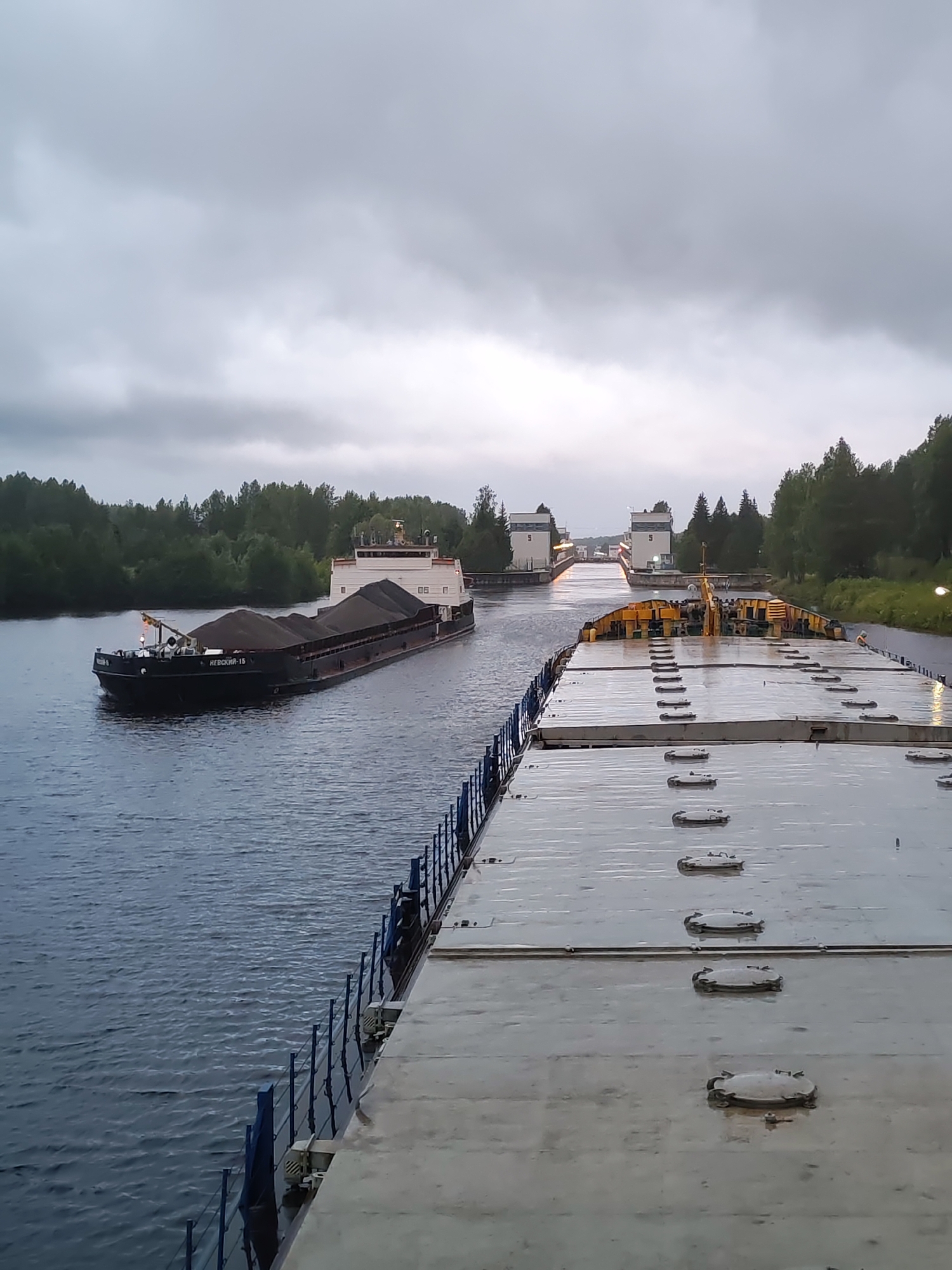 An overcast, gloomy day scene depicts a body of water with two barges. In the foreground, a barge with a flat, metallic deck is positioned alongside a darker body of water. The deck is wet and reflects the overcast sky, and it features multiple oval-shaped hatch covers arranged in rows. The barge is bordered by a blue metal railing. Further in the distance, a second barge, loaded with dark material that appears to be coal or gravel, is moving along the water. This barge has a more structured superstructure at its rear, with windows and a prominent white section. On the left side of the waterway, a dense line of green trees borders the scene, extending into the background. In the far distance, a series of white structures with multiple doors and windows are visible, suggesting a canal lock system. The overall atmosphere is muted due to the cloudy sky, with a slight sheen of water visible on surfaces, possibly indicating recent rain. The location is the city of Alekseevskoe, Russia.