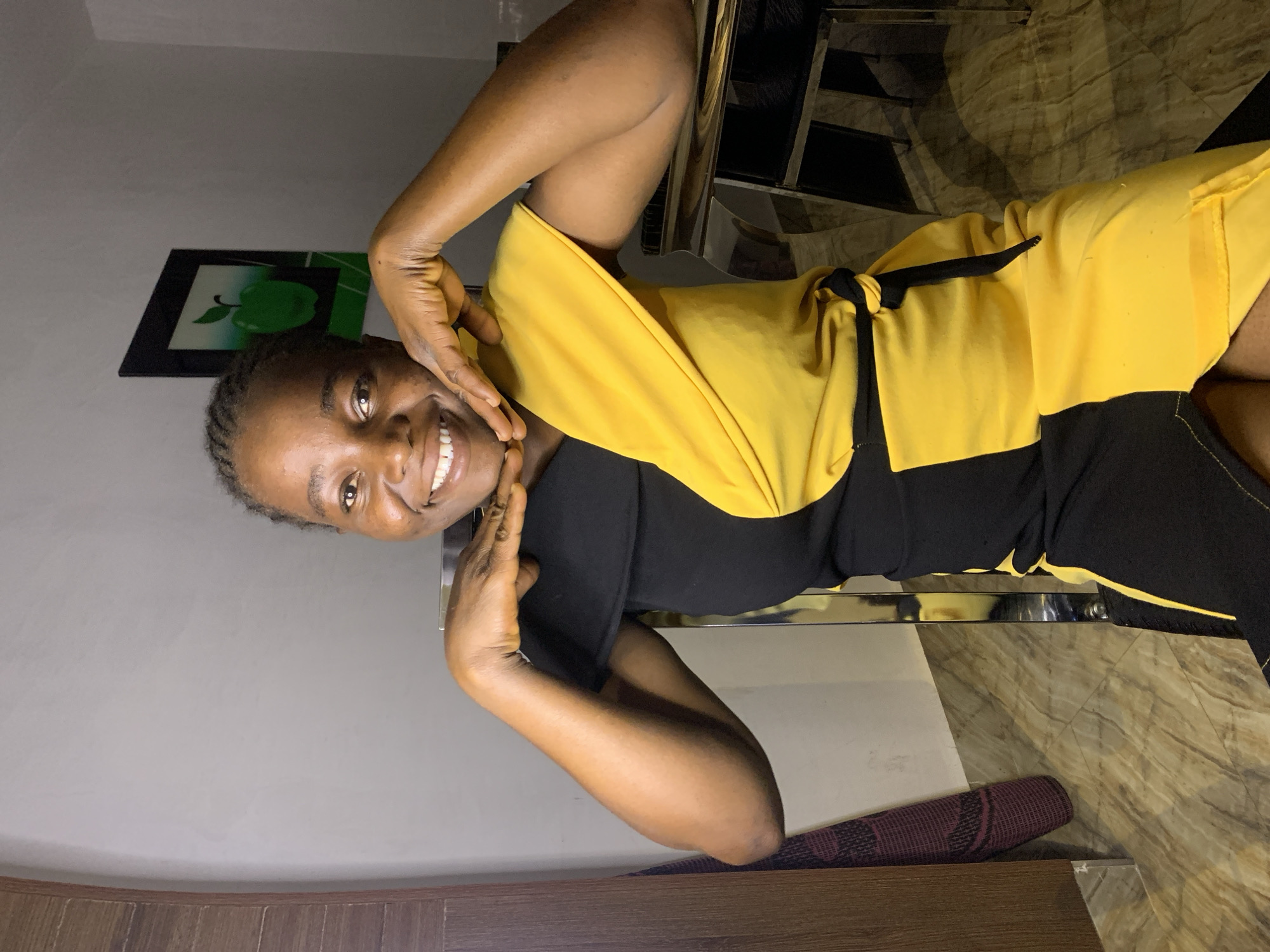 A young woman is posing for a photo indoors. She is wearing a black and yellow dress and has her hands on her face. A painting of a green apple is on the wall behind her. The location is possibly in Jos, Nigeria, but this is not definitively confirmed by the image.