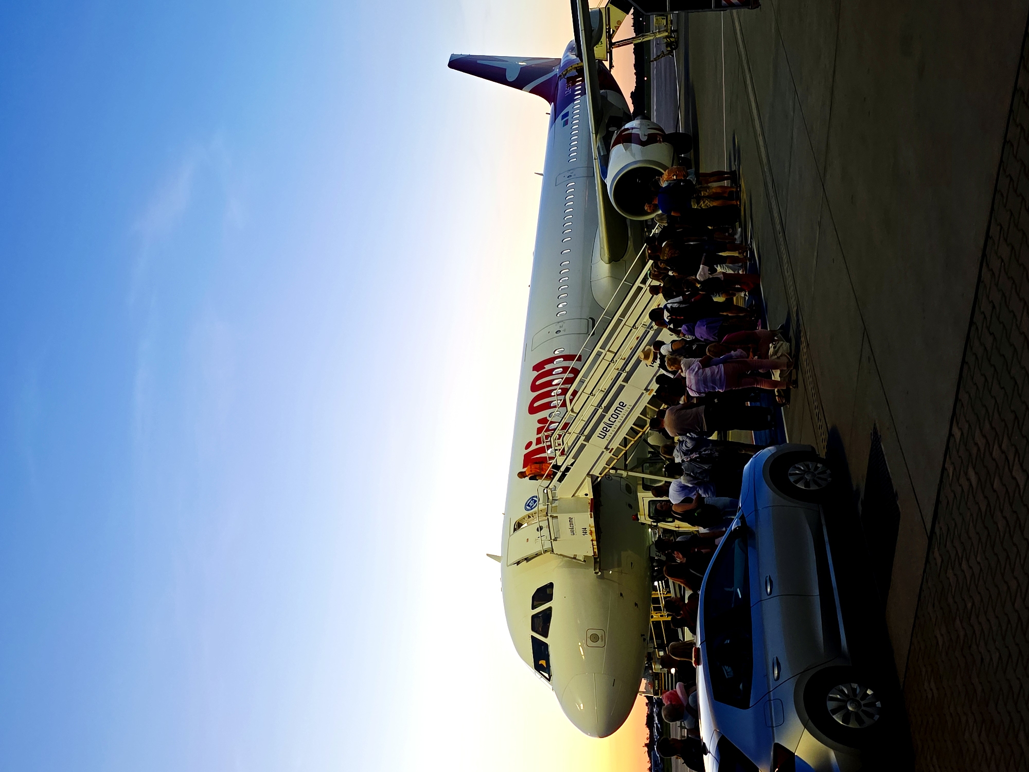 An overhead view of an airplane on an airport tarmac. The airplane is white with a red logo on the side and a purple tail fin. A set of stairs leads up to the plane's entrance. A crowd of people is boarding the plane. A silver car is parked in the foreground. The sky is blue with a hint of orange at the horizon, suggesting it is either sunrise or sunset. The scene appears to be at Poznań–Ławica Airport, based on the characteristic architecture and the presence of a Wizz Air airplane.
