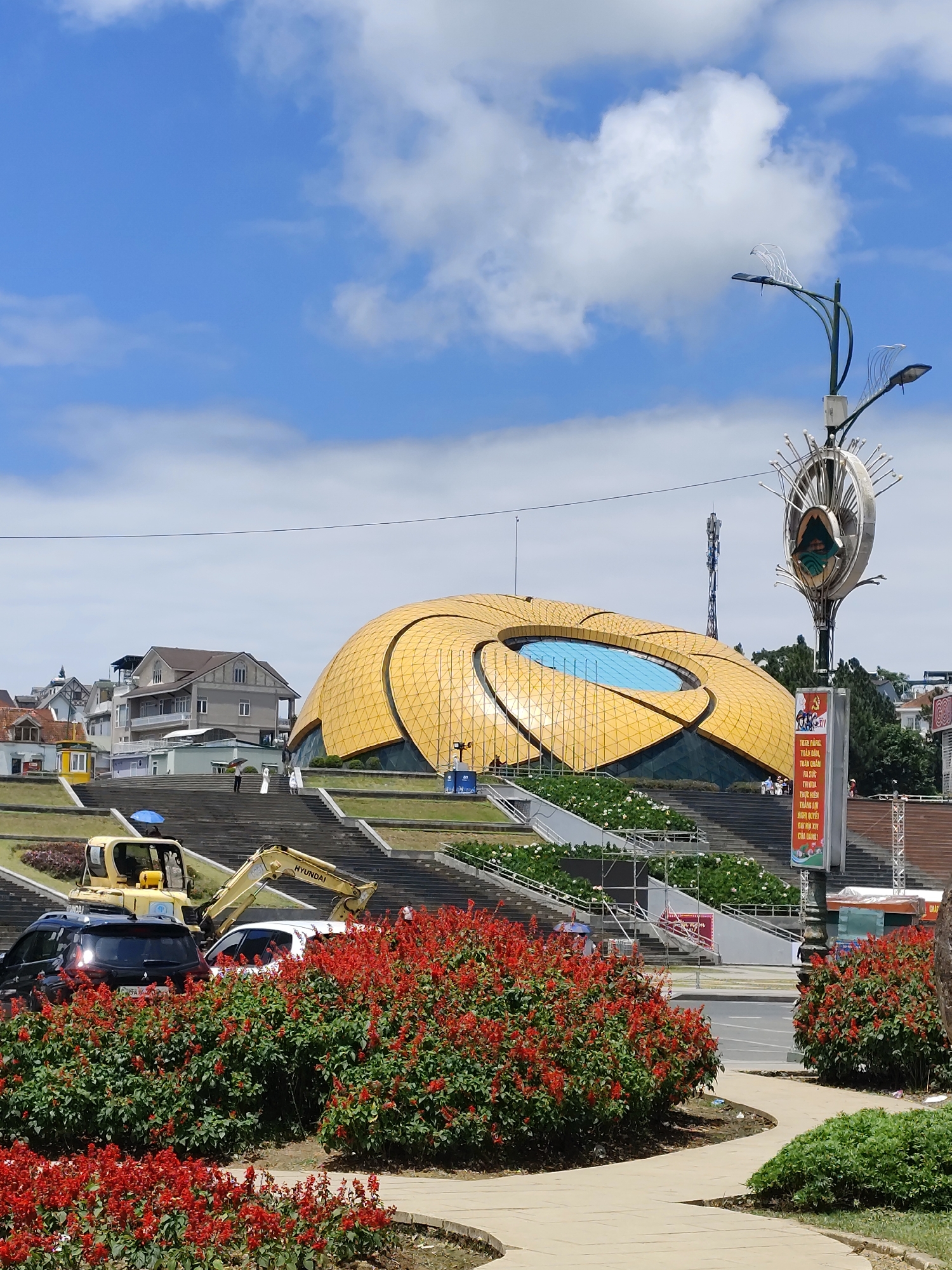 In Da Lat, Vietnam, the image captures a sunny day with a bright blue sky and scattered white clouds. A large, distinctive yellow dome-shaped building, likely a cultural center or performance venue, dominates the background. Its segmented, petal-like exterior and a central blue circular window are visible. In front of the building, a wide set of concrete stairs leads up to the entrance. Several people are walking up the stairs, some holding umbrellas. Construction equipment, including a yellow Hyundai excavator, is positioned on the stairs, indicating ongoing work or maintenance. To the left of the stairs, a row of houses with various architectural styles can be seen. In the foreground, lush green bushes are interspersed with vibrant red flowers, bordering a light-colored paved pathway. A modern lamppost with an artistic design stands to the right of the pathway, with a banner attached to its pole featuring Vietnamese text and an illustration. Cars are parked near the bottom of the stairs, partially obscured by the foliage. The overall scene depicts a public space in Da Lat, showcasing a blend of architecture, nature, and urban activity.