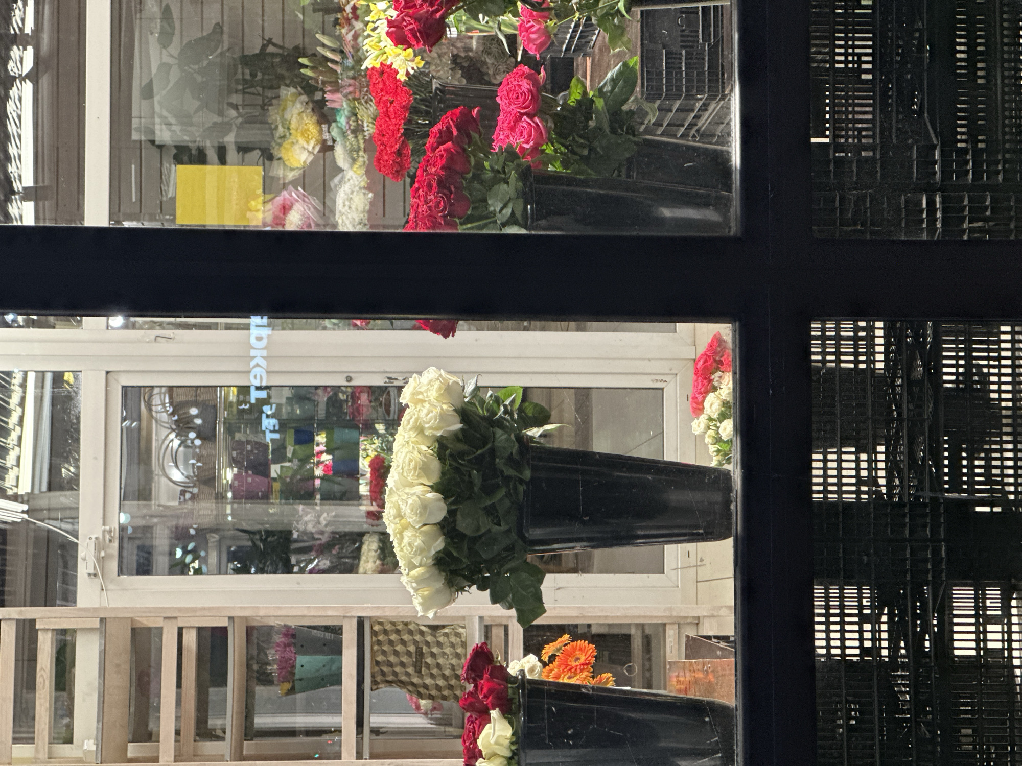 A street-level view, taken through a window frame, captures a flower shop's interior, possibly in Chaikovsky, Russia, although direct confirmation is absent. Various bouquets, including red and white roses, are arranged in black vases. A ladder stands near the window, obscuring some of the shop's contents. The reflection on the glass adds to the scene's complexity, revealing more flowers and what appears to be the interior. Part of what appears to be an air conditioning unit appears in the right of the image.