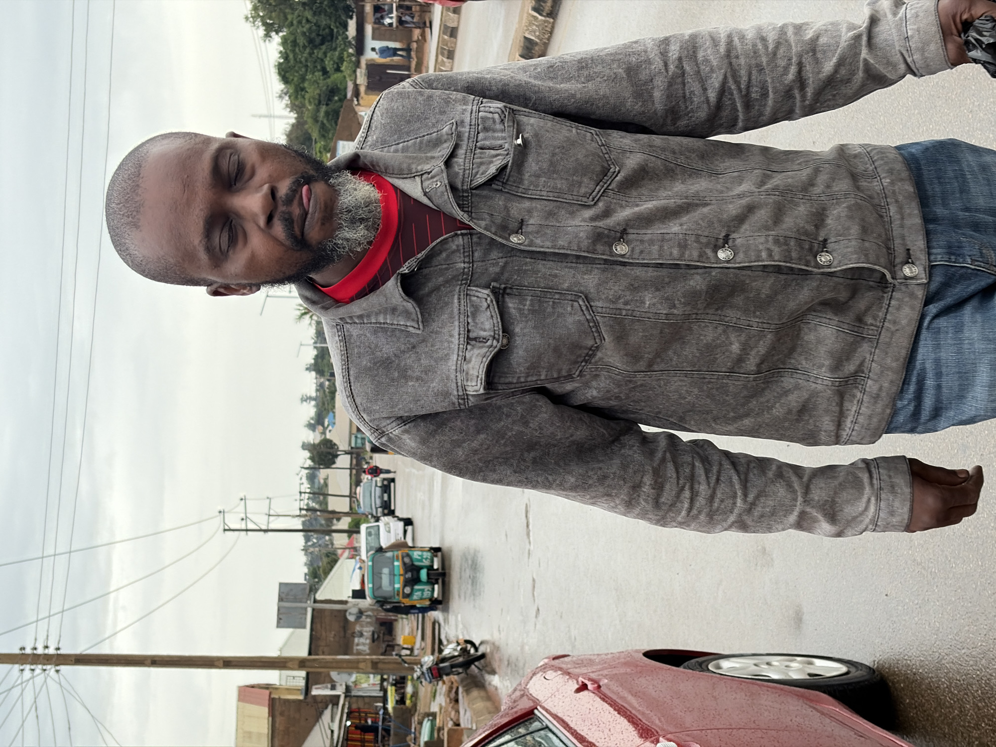 A man with a gray beard and closed eyes walks down a wet, paved street in Jos, Nigeria. He is wearing a gray denim jacket over a red and black striped shirt and blue jeans. He is holding a black plastic bag in his right hand. The street is lined with buildings and trees, and there are several vehicles, including a tuk-tuk, a motorcycle, and cars, on the road. The sky is overcast.