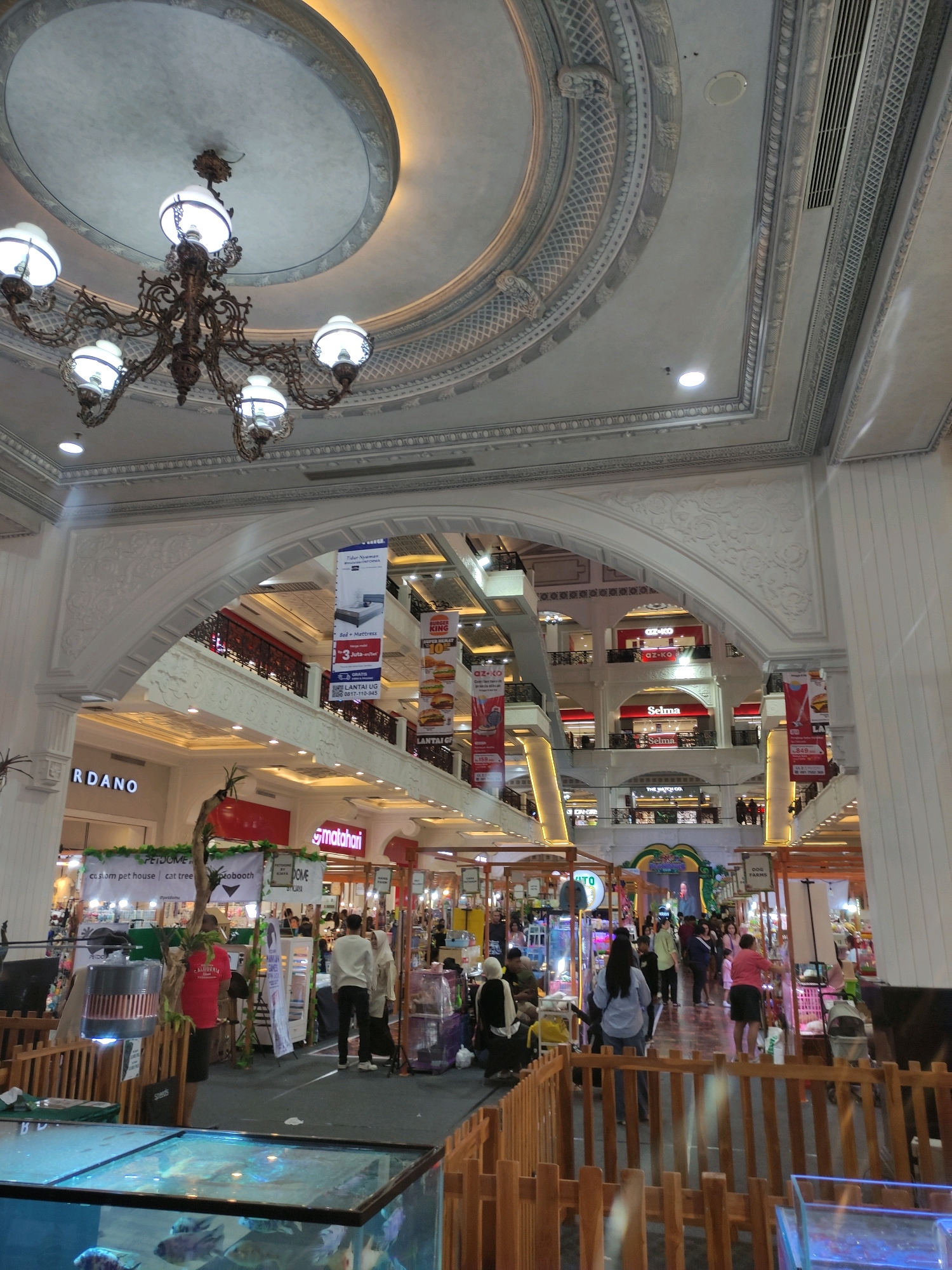 The image captures the interior of a multi-level shopping mall, likely located in Sinduadi, Indonesia, as indicated by the presence of "Matahari" department store branding. The scene is brightly lit, with a large, ornate chandelier hanging from the ceiling. The architecture features grand arches, decorative moldings, and multiple levels of walkways and storefronts. People are visible on different levels, browsing shops and walking along the corridors. Banners advertising various products and services, including "Burger King" and "Selma" furniture, are displayed throughout the mall. In the foreground, a wooden fence encloses an area with an aquarium containing fish, suggesting a retail space for aquatic pets or a decorative display. The overall impression is one of a bustling commercial environment.