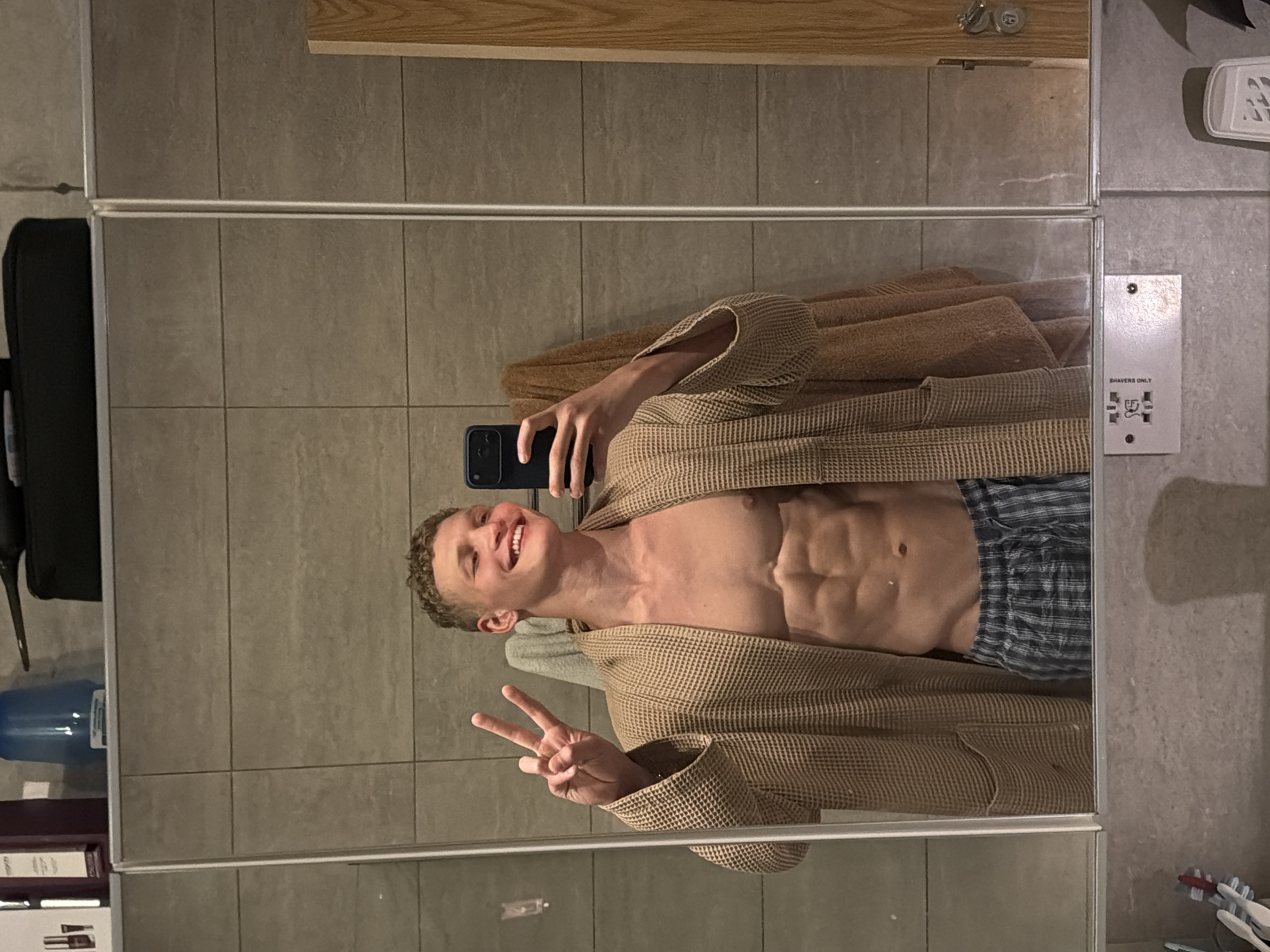 A person is reflected in a mirrored wall wearing a waffle-weave bathrobe and plaid shorts. They are holding a smartphone, appearing to take a selfie, and making a peace sign with their left hand. The person has light skin, short curly hair, and is smiling. Their abdomen is visible, showing well-defined abdominal muscles. The background includes bathroom tiles, a cabinet with toiletries, a suitcase, and a water cooler. A shaver outlet is visible on the wall. The location is not discernible within the city of London.